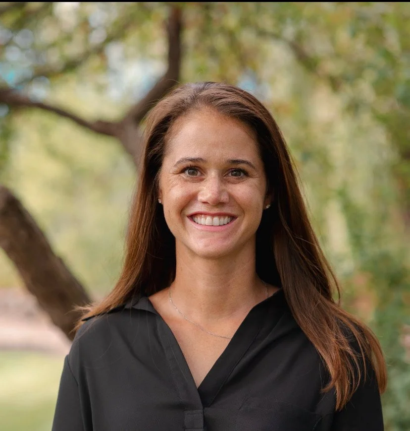 A smiling woman with long brown hair, wearing a black top, standing outdoors in front of a tree with green and yellow leaves.