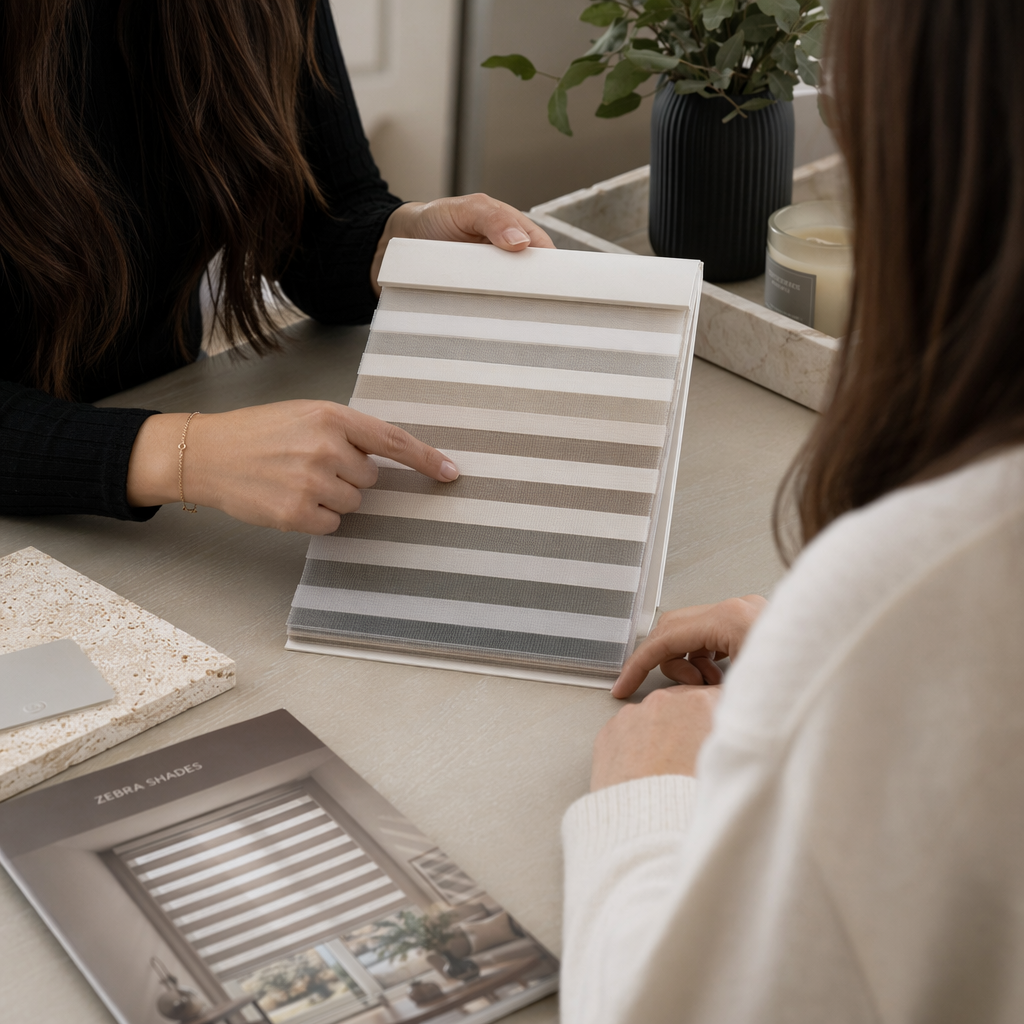 Two women discussing fabric or paint swatches at a table, with a book titled 'Zebra Shades' and a potted plant in the background.
