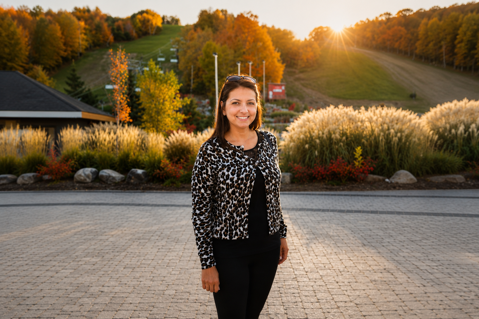 A smiling woman in a leopard print jacket and black top standing outdoors on a cobblestone surface during sunset with fall foliage and hills in the background.