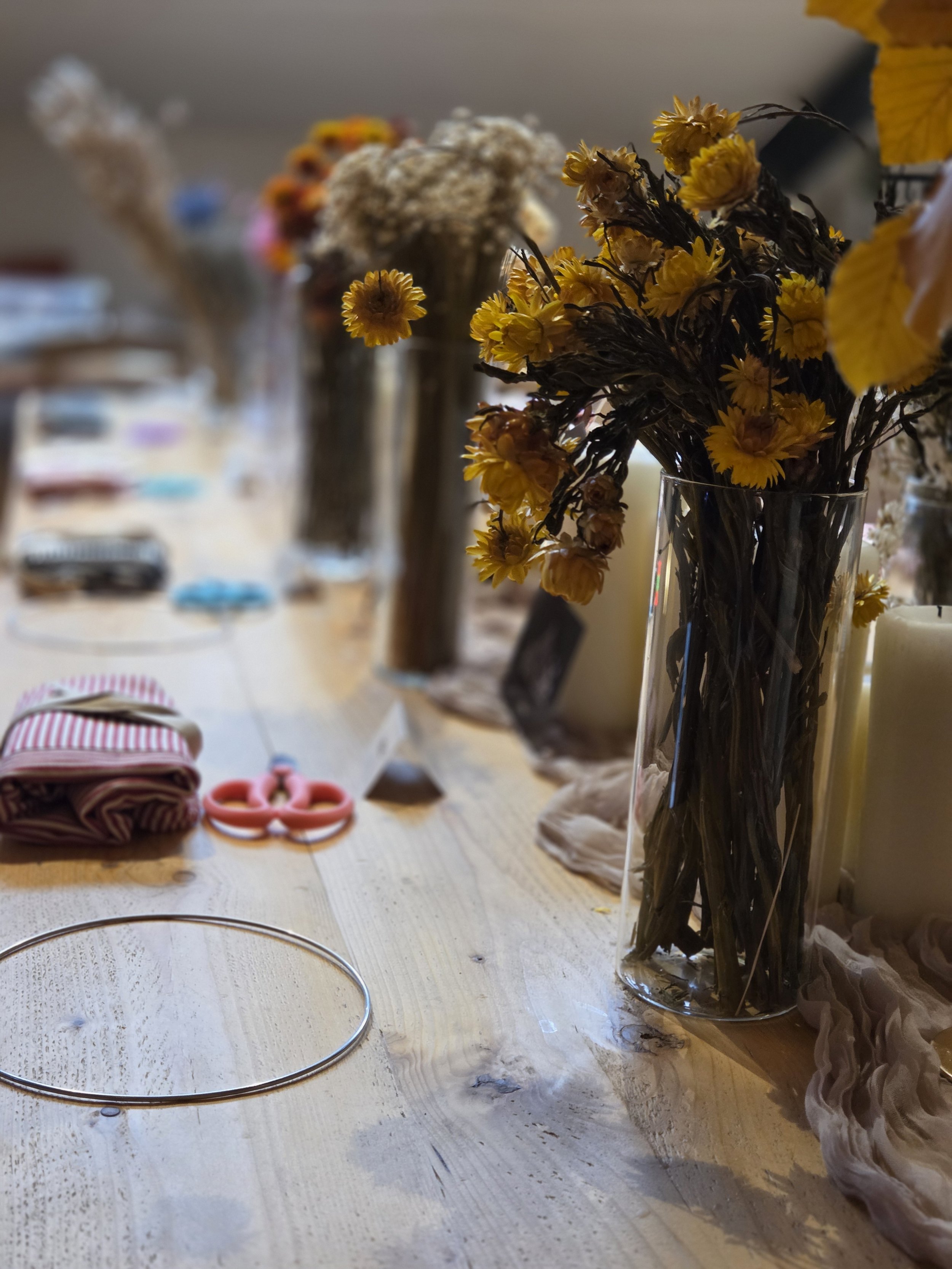 A bouquet of yellow and white dried flowers in a tall, clear glass vase on a wooden table, with other vases and objects blurred in the background.
