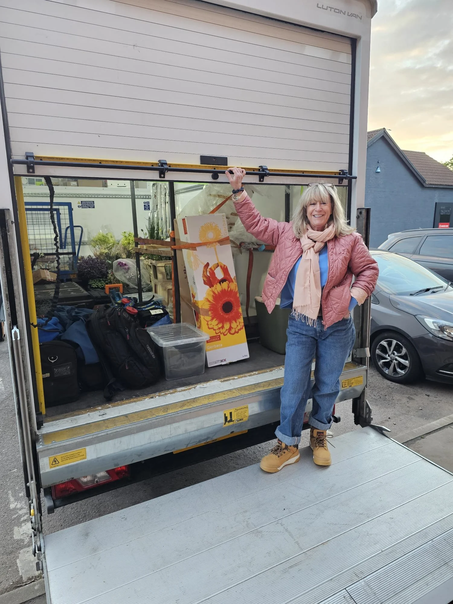 Woman smiling and standing on a loading dock next to an open truck filled with bags, boxes, and gardening supplies during daytime.