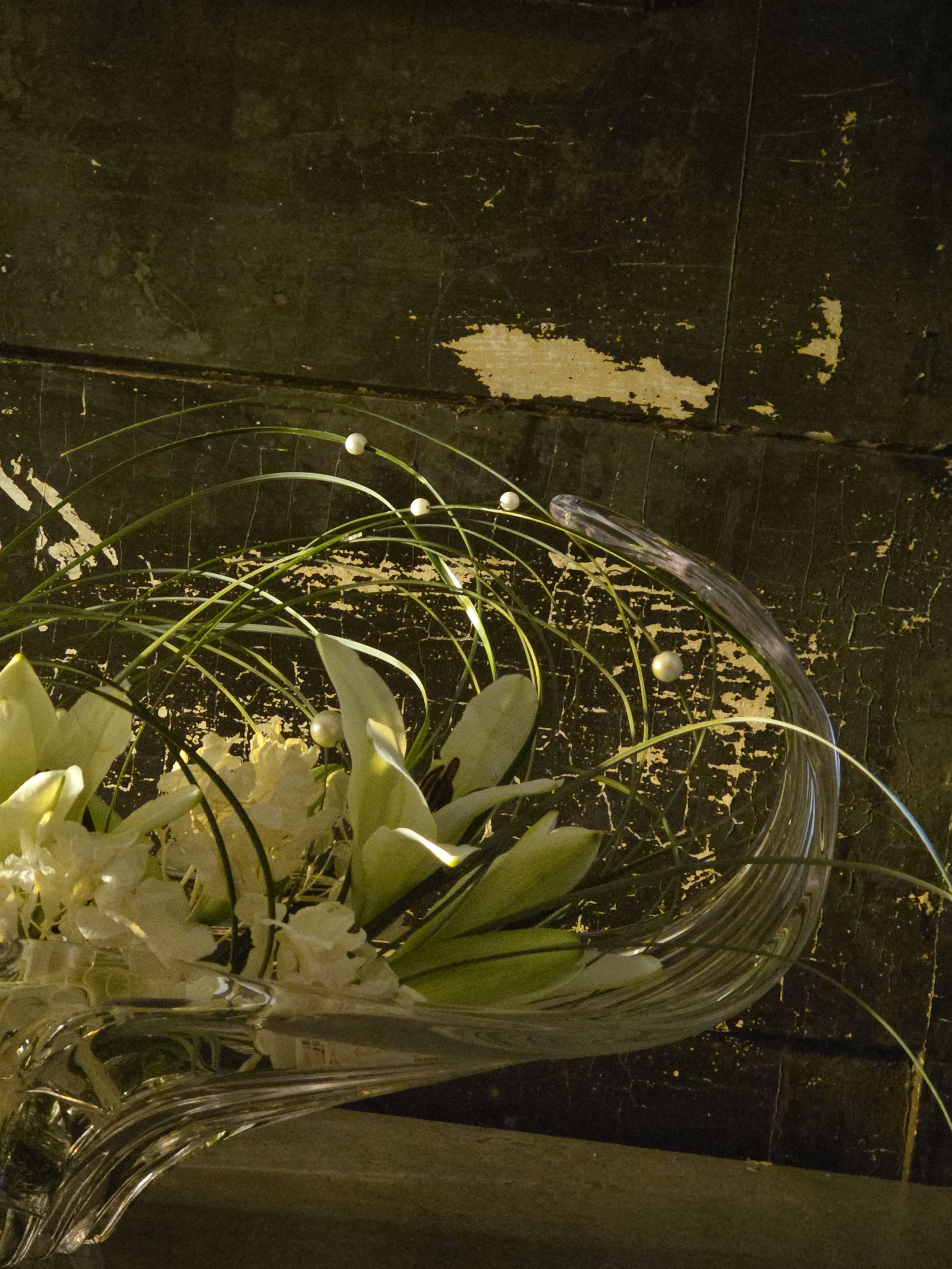 A glass bowl with white flowers and decorative white balls, placed on a dark surface, with a black background that has peeling paint and scratches.