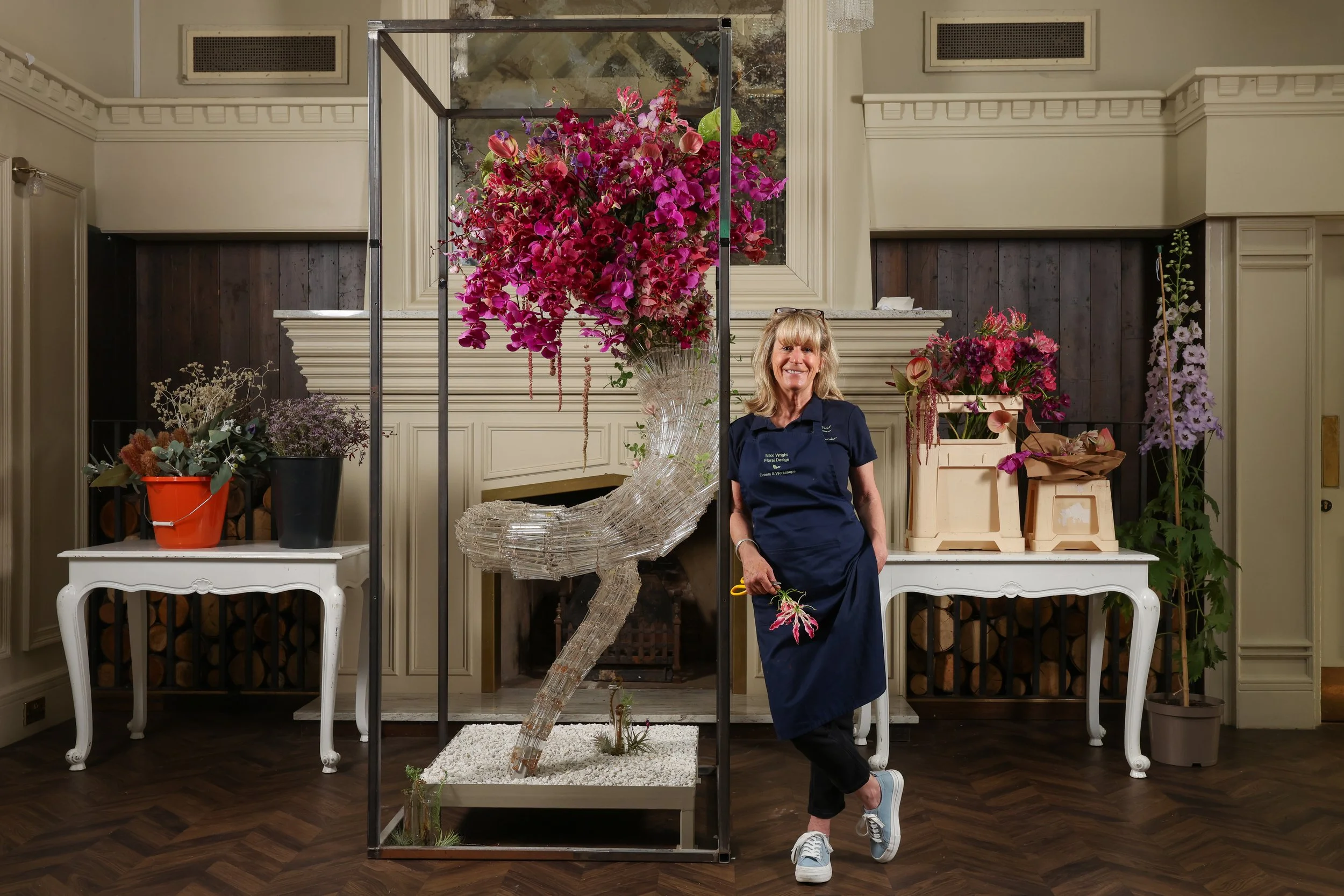A woman standing next to a large floral arrangement and a glass sculpture in a room with cream-colored walls and dark wood flooring.