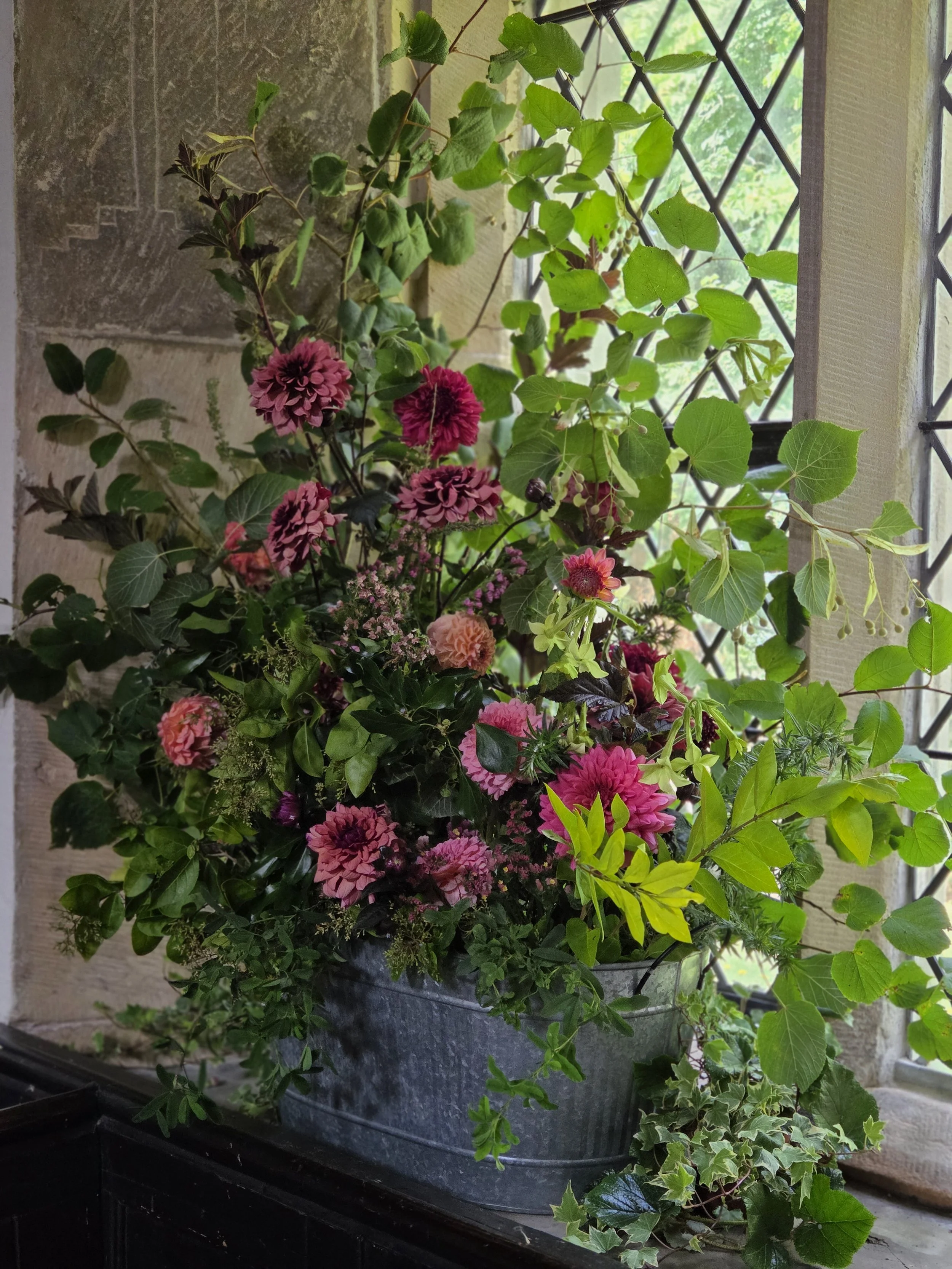 A large metal planter filled with pink and purple flowers, green leafy plants, and trailing ivy, situated on a black surface near a stone wall and a window with a metal grid, sunlight illuminating the plants.