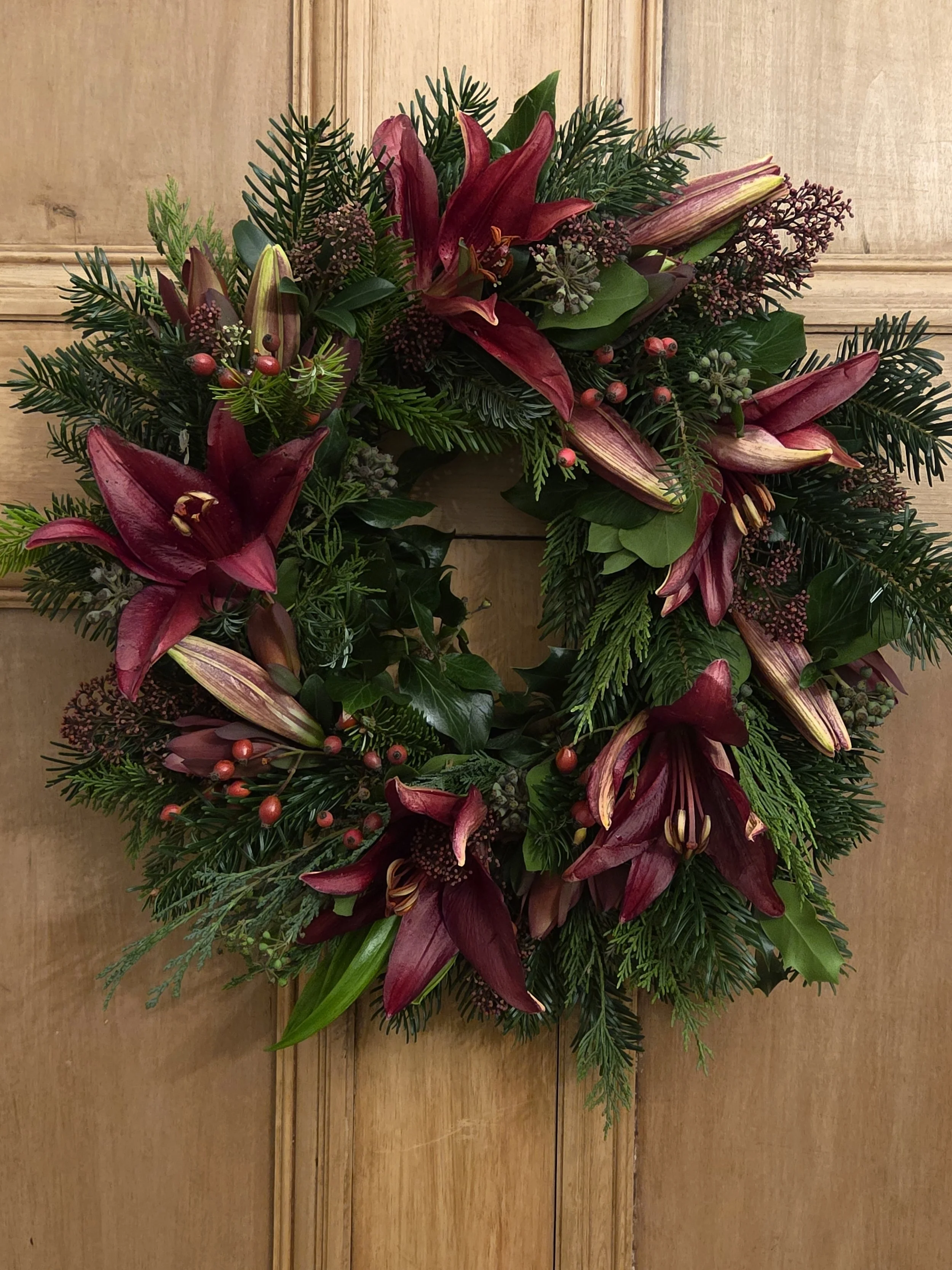 A Christmas wreath decorated with red lilies, pine branches, holly leaves with red berries, and other greenery, hanging on a wooden wall.