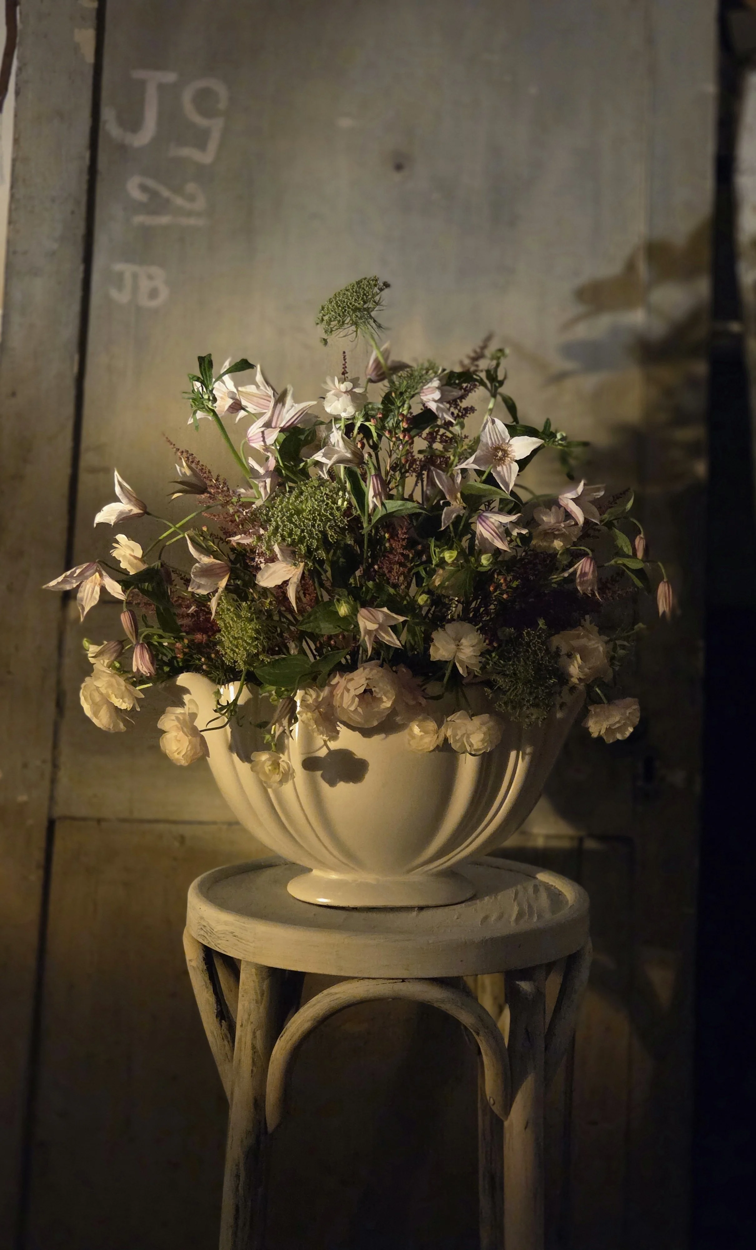 A ceramic vase with a floral arrangement of white and pink flowers, greenery, and small buds, placed on a round wooden stand in a rustic setting with textured wall background.
