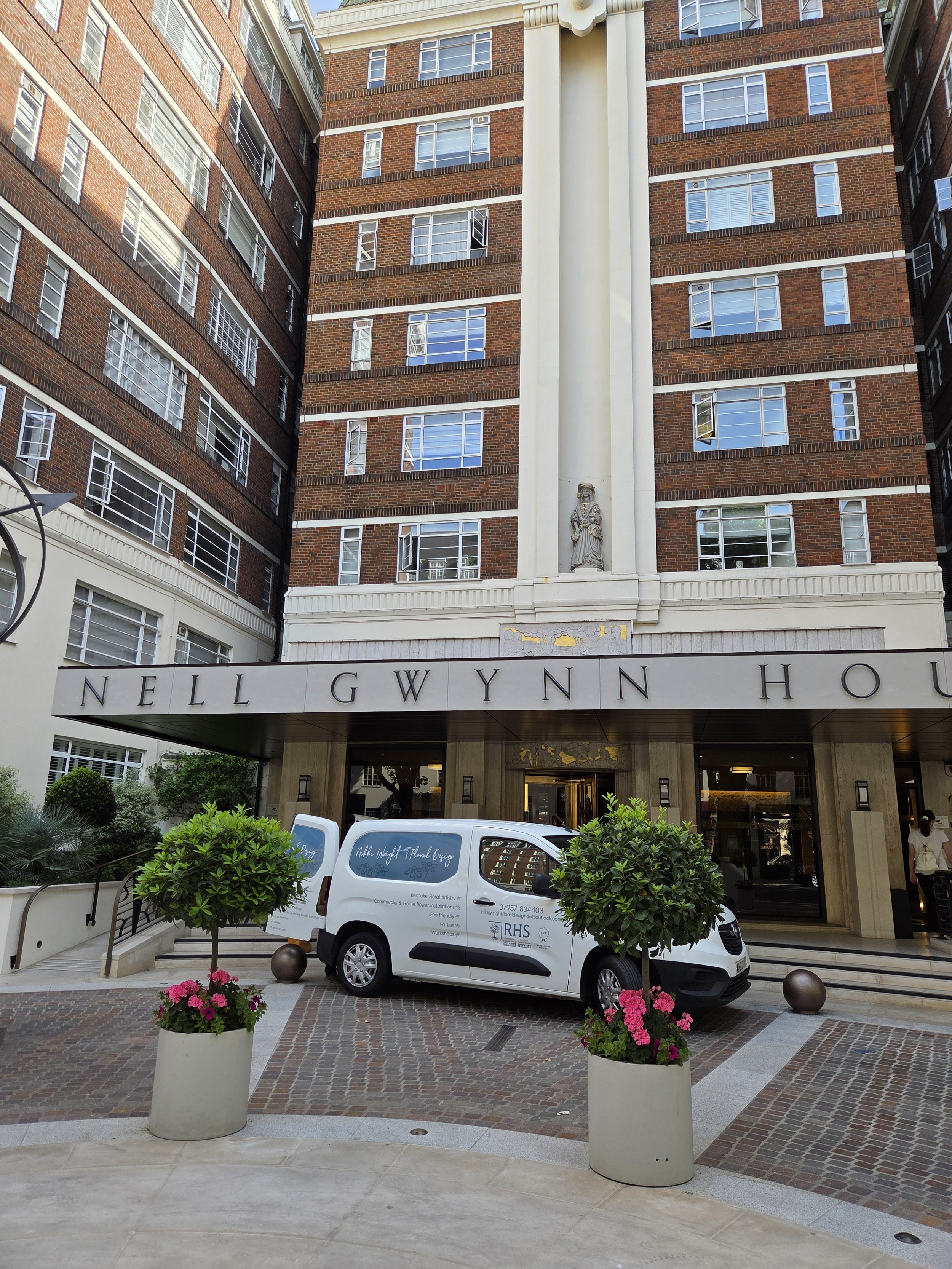 The image shows the entrance of a building with tall brick walls, several open and closed windows, and a statue above the doorway. There is a white van parked in front of the entrance, with some pink flowers in pots on either side of the parking area