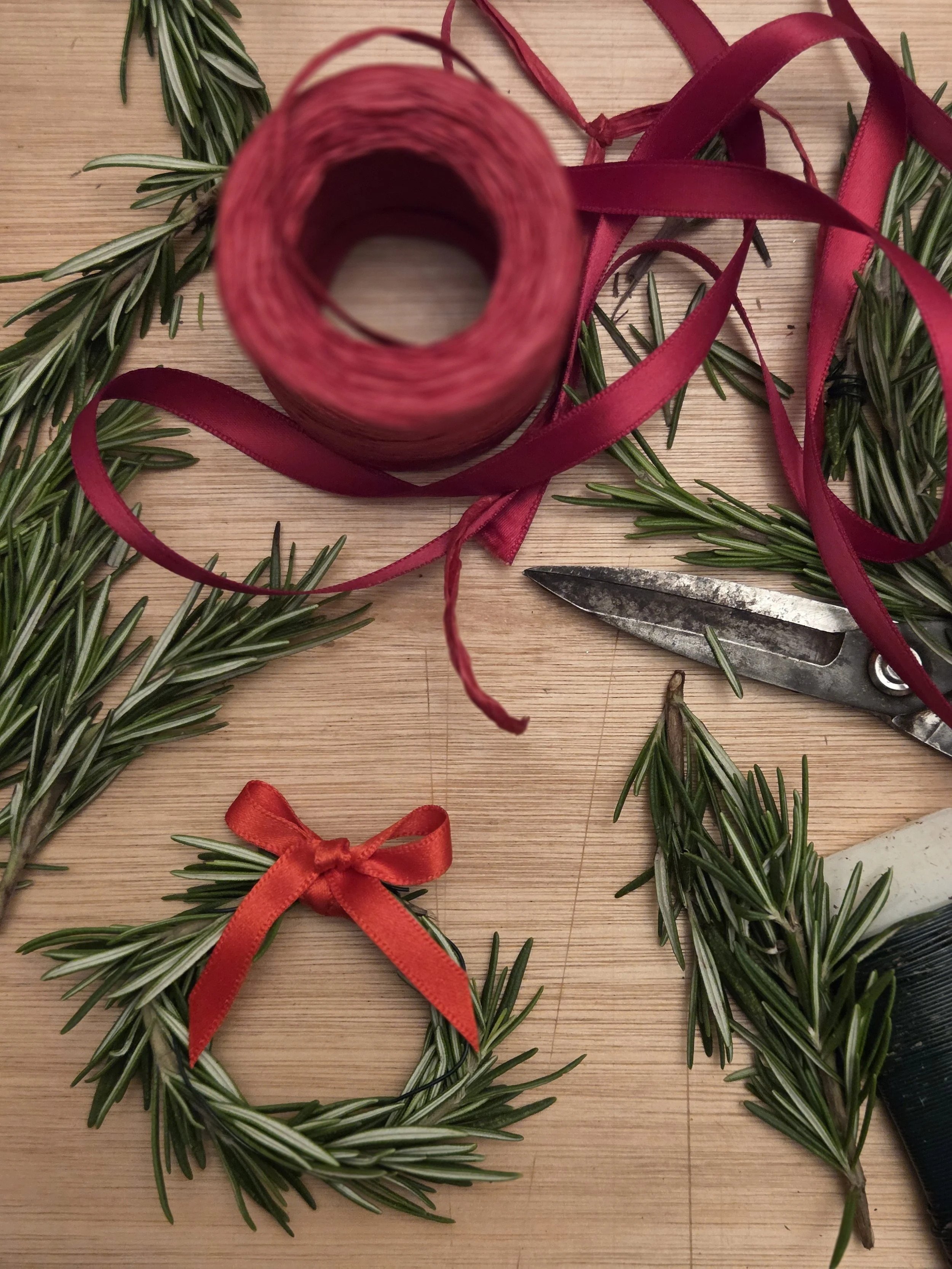 Green pine branches with some red ribbon bows, a spool of red ribbon, and a pair of scissors on a wooden surface.