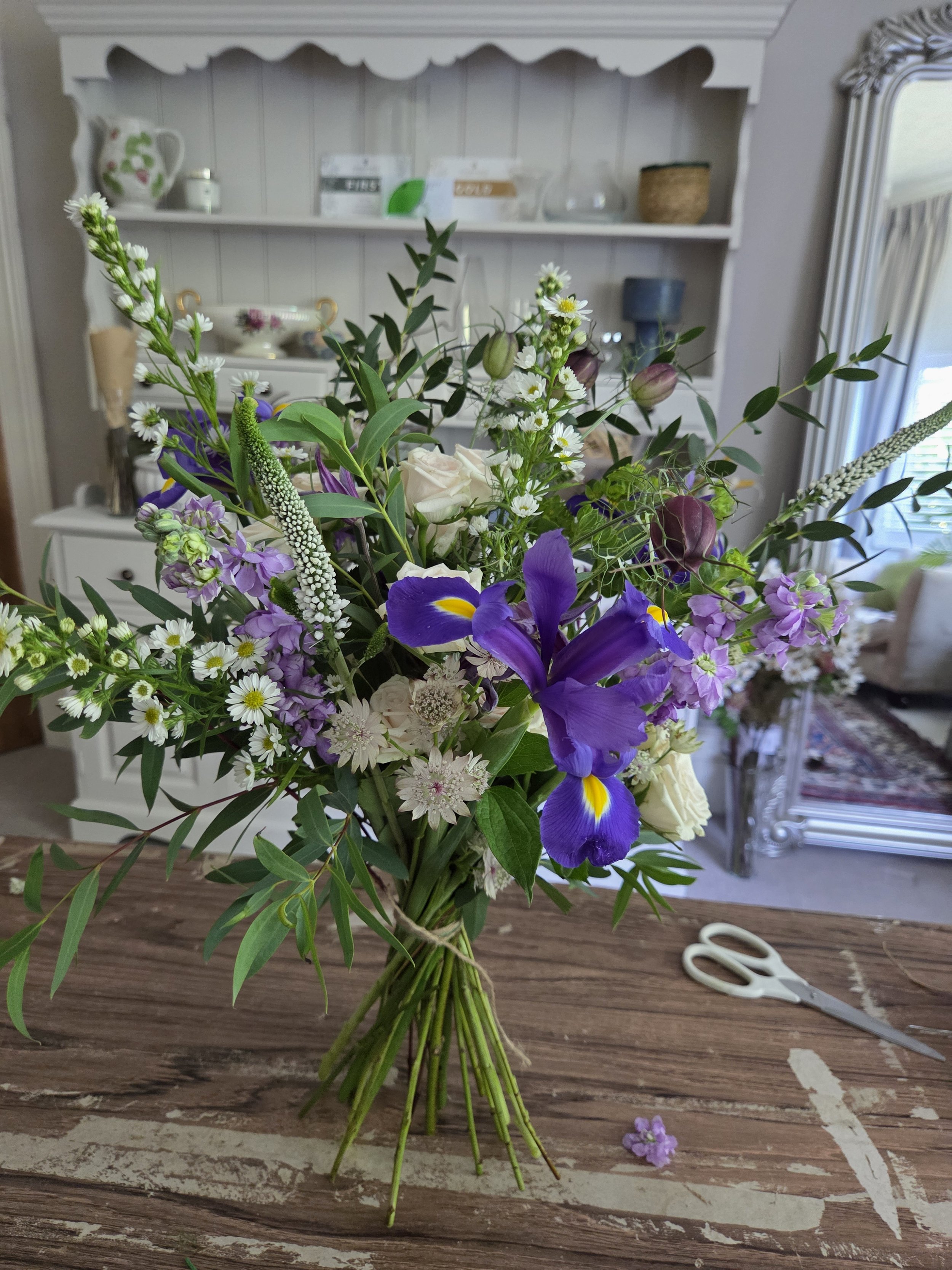 A bouquet of various flowers, including purple irises, white daisies, and other mixed wildflowers, sits on a wooden table with a pair of white scissors nearby. The background features a white wooden shelf with decorative items and a mirror to the rig