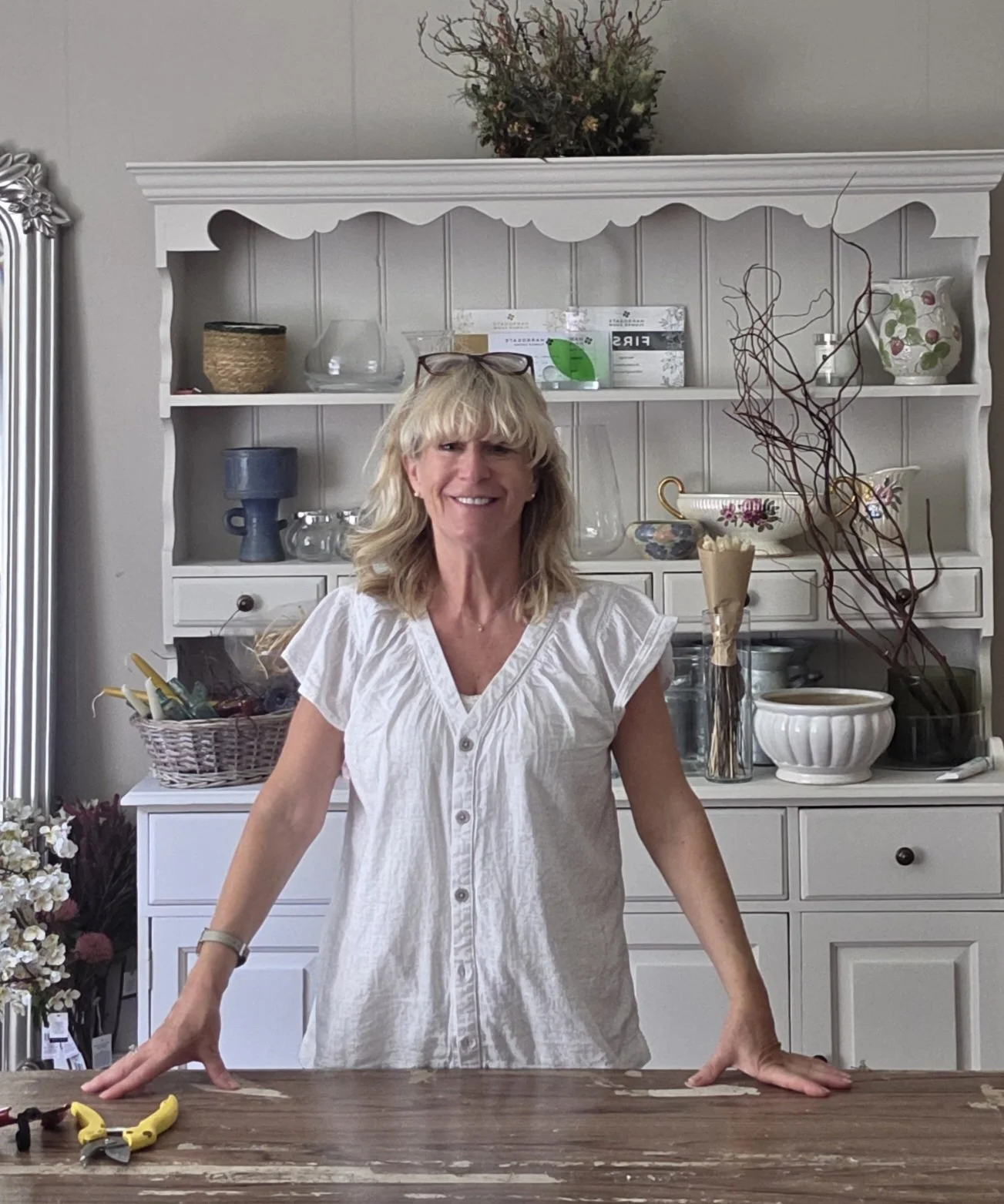 A smiling woman with blonde hair and glasses resting on her head stands behind a wooden table in a bright, decorated room. The room has white shelves filled with various ceramics, glassware, and decorative items. There are flowers and branches as decor, with a large vase and a basket of flowers on the side.