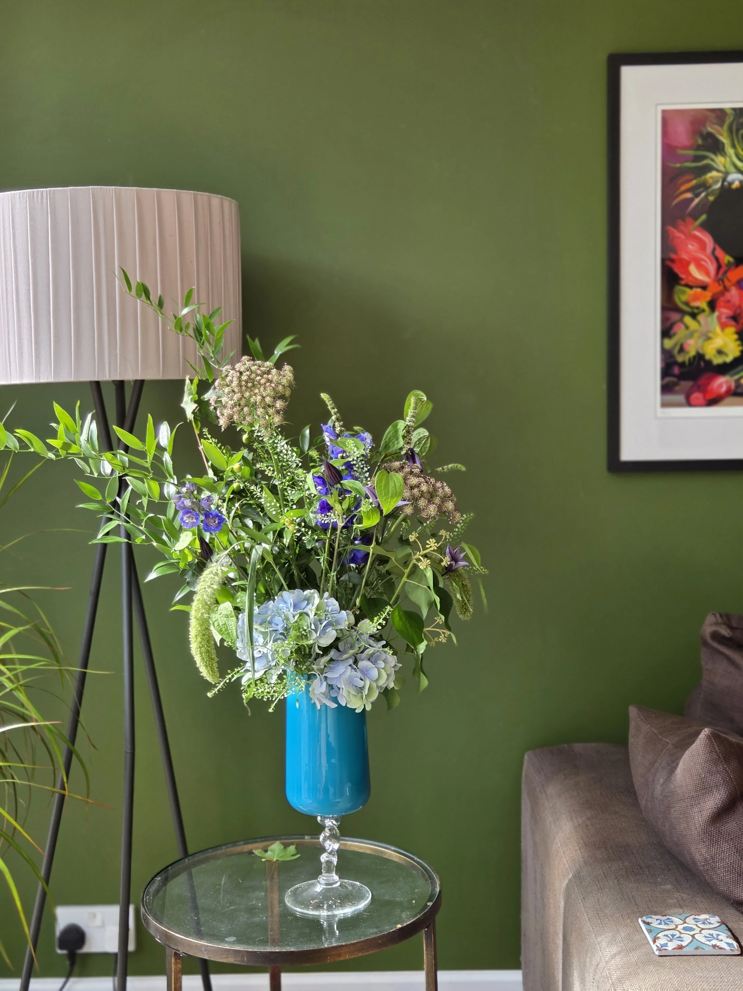 A green wall with a framed colorful floral painting, a floor lamp, a glass table with a tall blue vase holding a mixed bouquet of flowers and greenery, and a beige sofa with a patterned booklet on it.