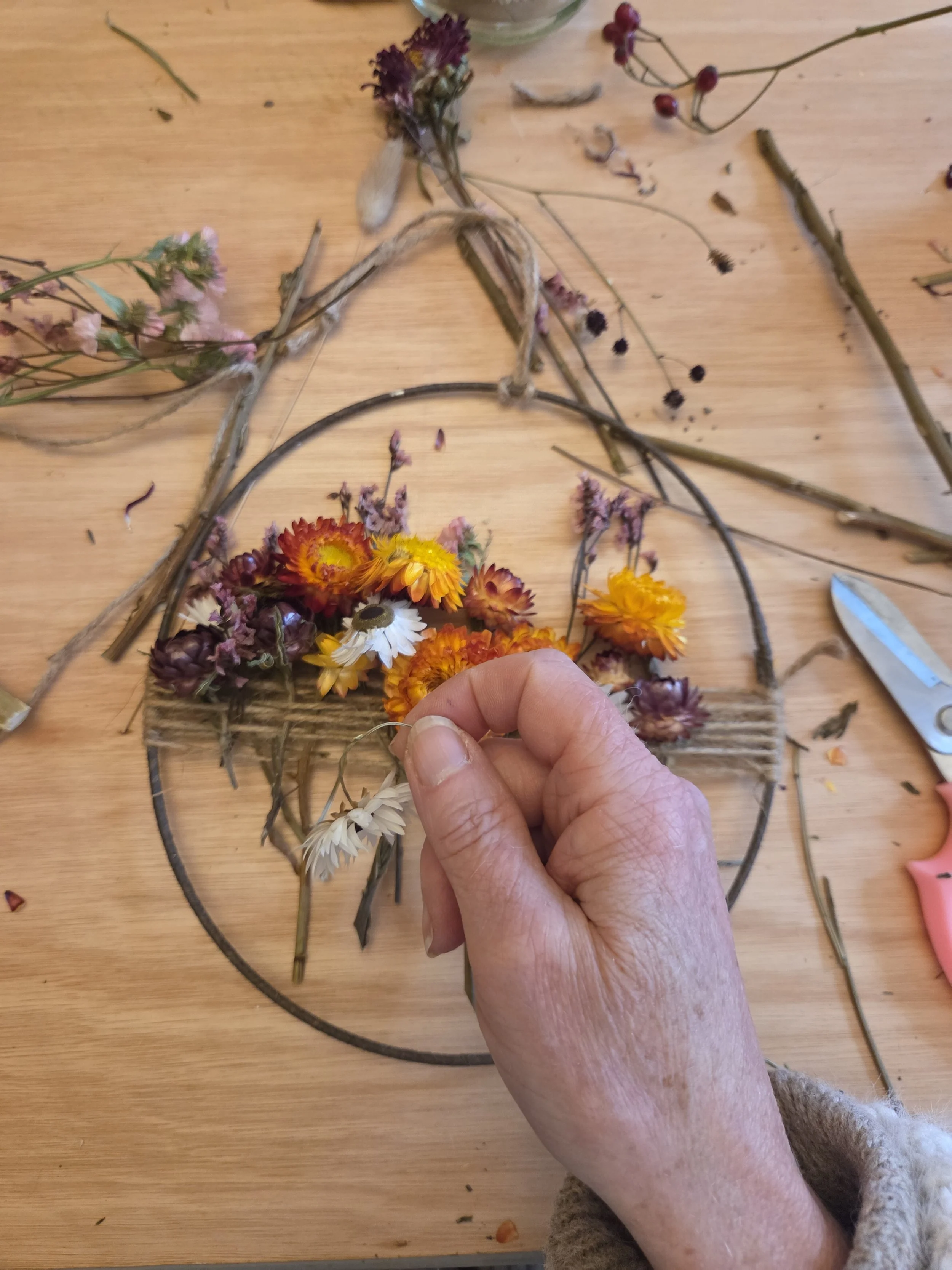 A person's hand arranging dried and fresh flowers in a circular wreath on a wooden table.