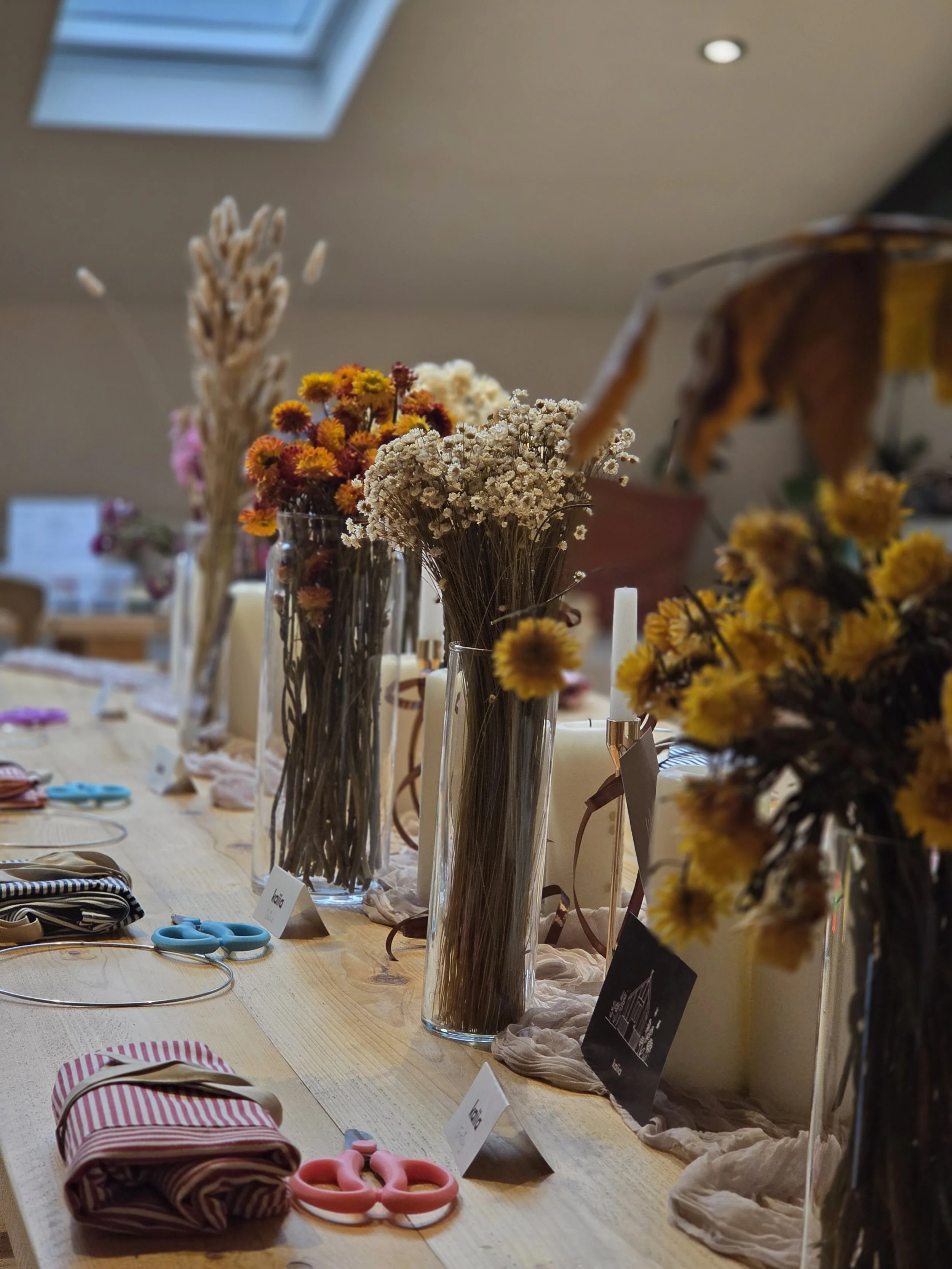 A table displaying vases of dried flowers, scissors, and handmade items, with labels and tags in a store or craft setting.