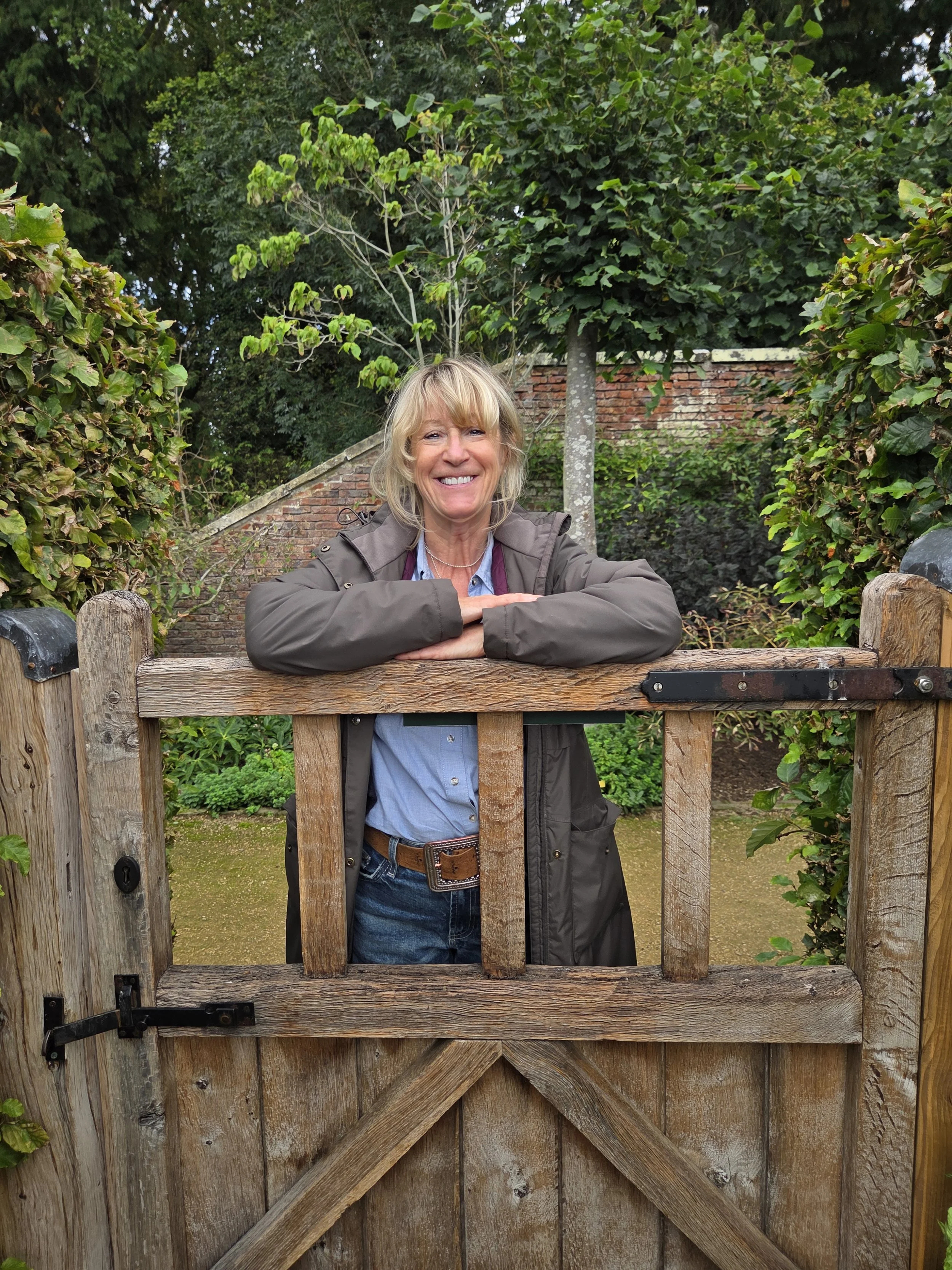 Nikki with blonde hair smiling and leaning on a wooden gate in a garden with trees and brick walls.