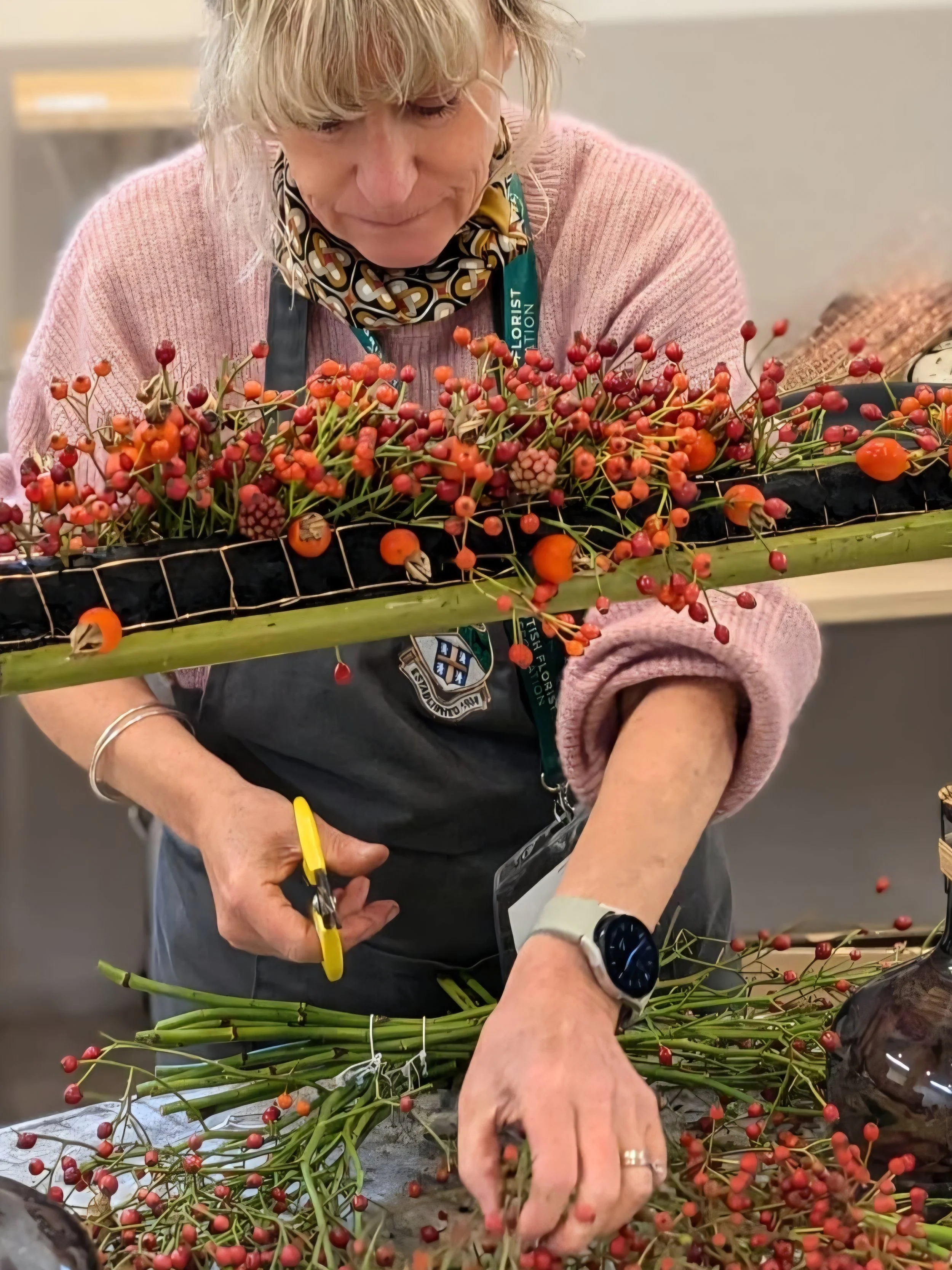 A woman in a pink sweater and patterned scarf is arranging red and orange berries and green stems on a table during a floral arrangement activity.