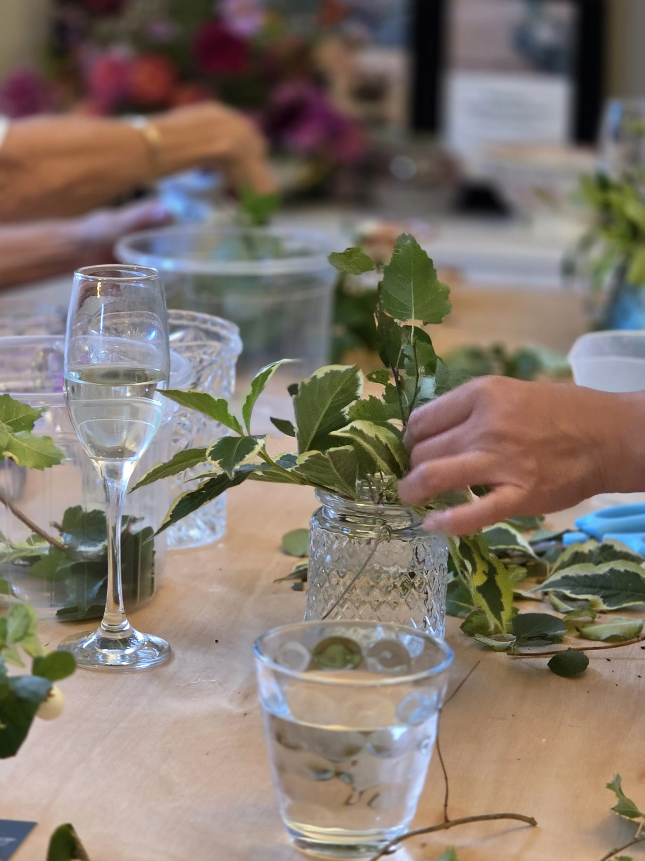 A person arranging green leaves in a glass vase on a table with glasses and other arrangements. In the background, a blurred person is working at the table.