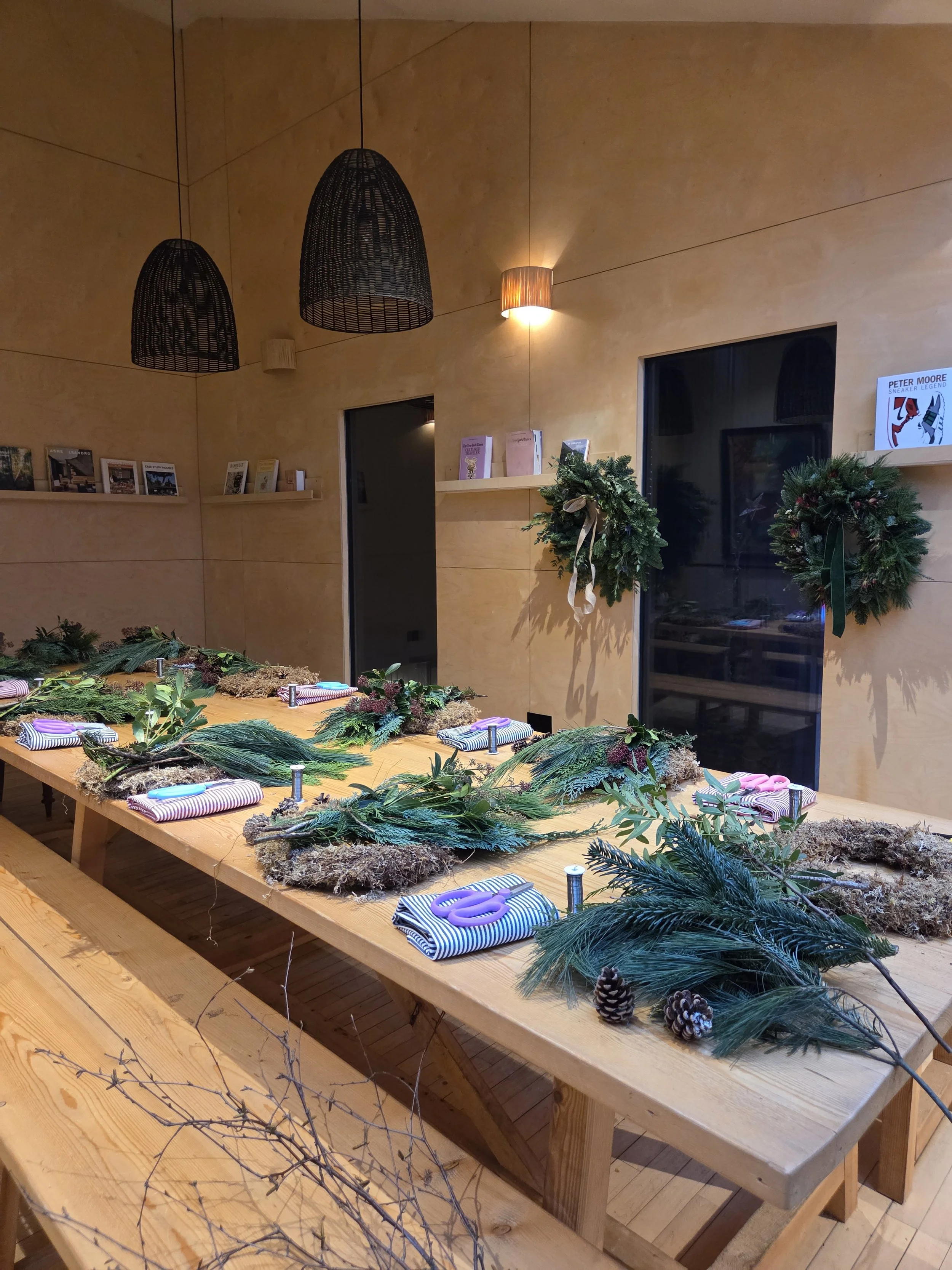 A long wooden table decorated with Christmas greenery, pine cones, and scissors, set for a holiday craft activity inside a room with beige walls, hanging pendant lights, and wreaths on the wall.