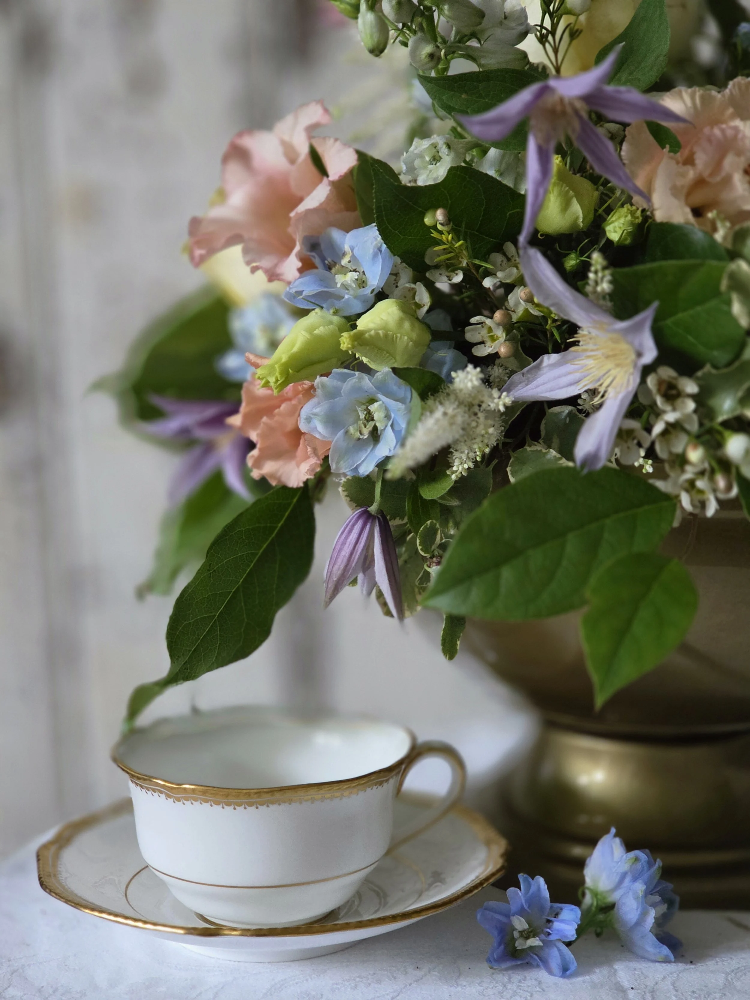 A floral arrangement with pastel pink, blue, and purple flowers in a white china teacup with gold trim, placed on a matching saucer, set on a white surface with additional flowers around.