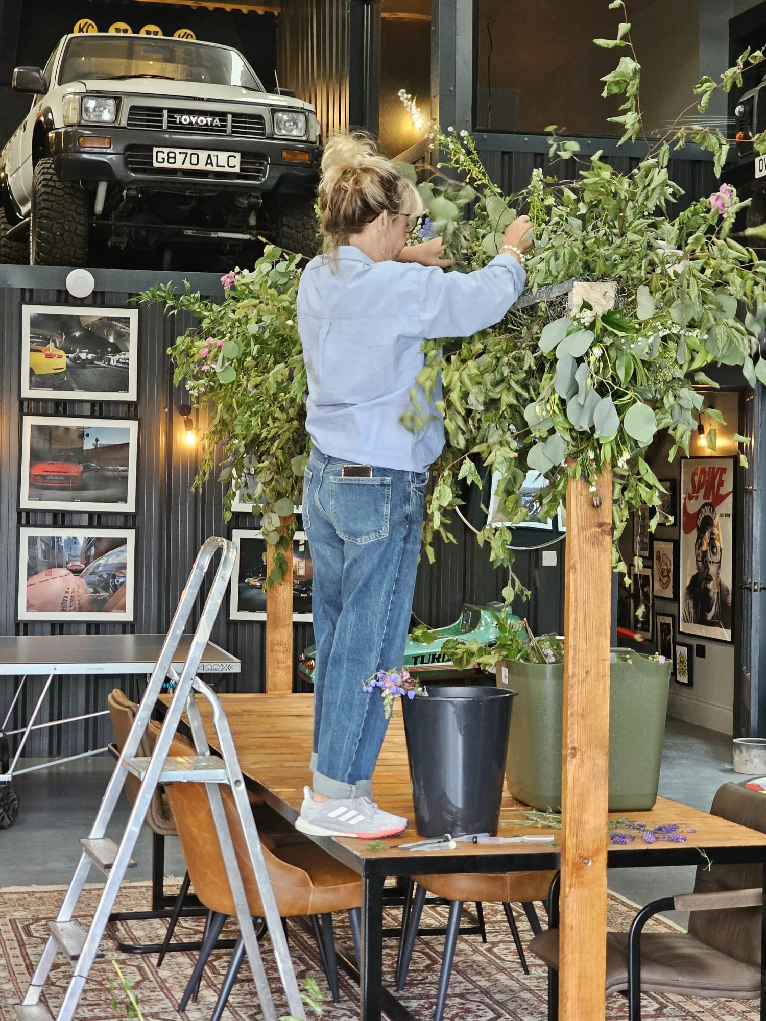 Nikki placing flowers in a large black pot behind a wooden frame and a small wooden platform. The scene appears to be indoors with car photos on the wall and a vintage car on display.