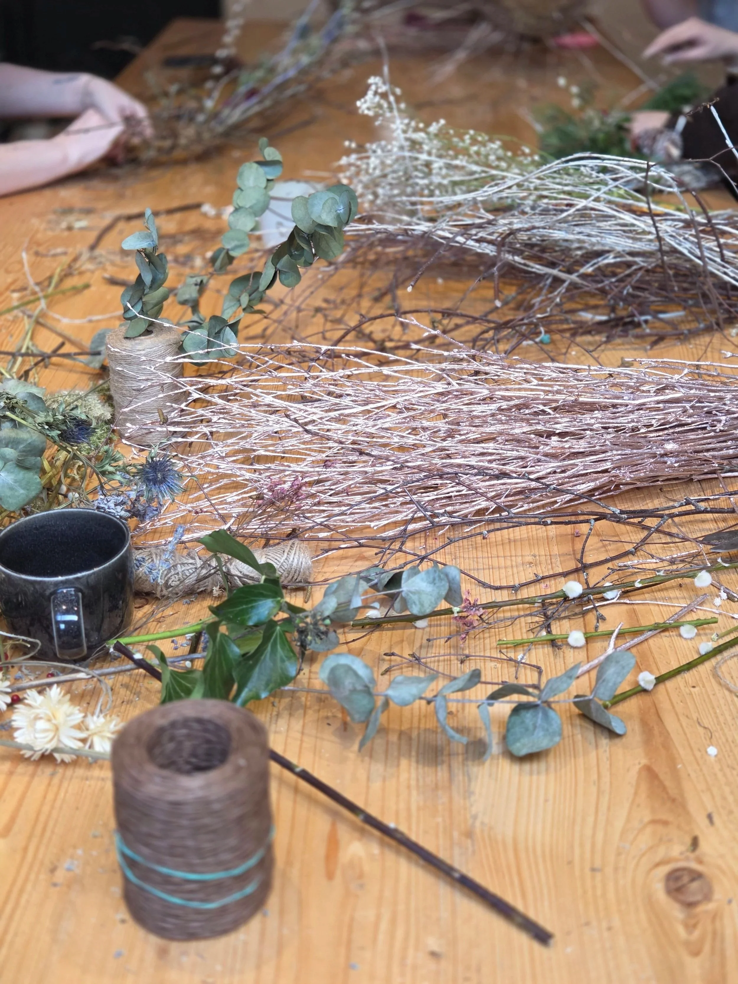 Various dried flowers, branches, and greenery arranged on a wooden table, with a spool of string and a black mug also visible.