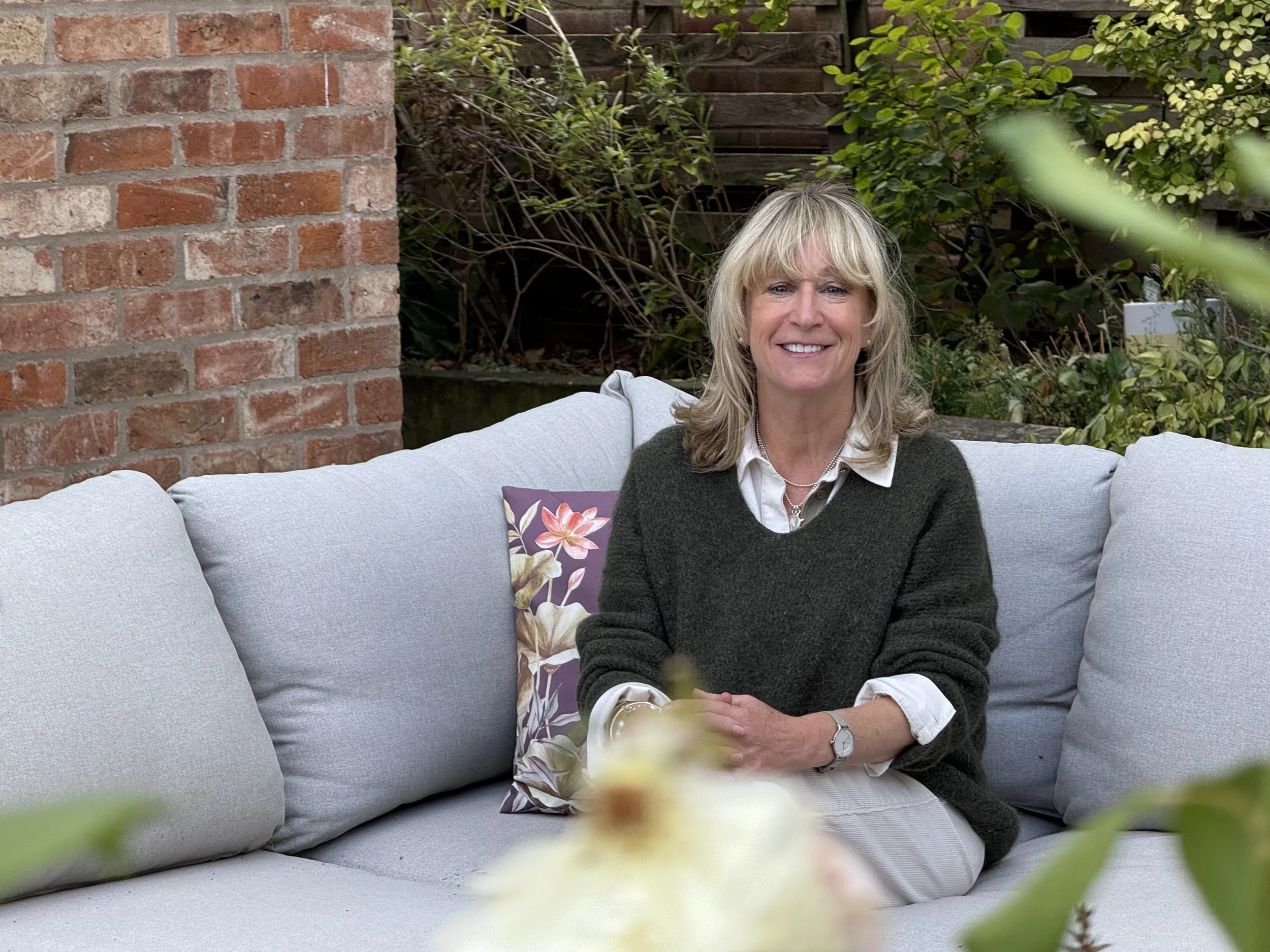 A woman with blonde hair, smiling, sitting outdoors on a gray patio sofa with a floral pillow, surrounded by garden plants.