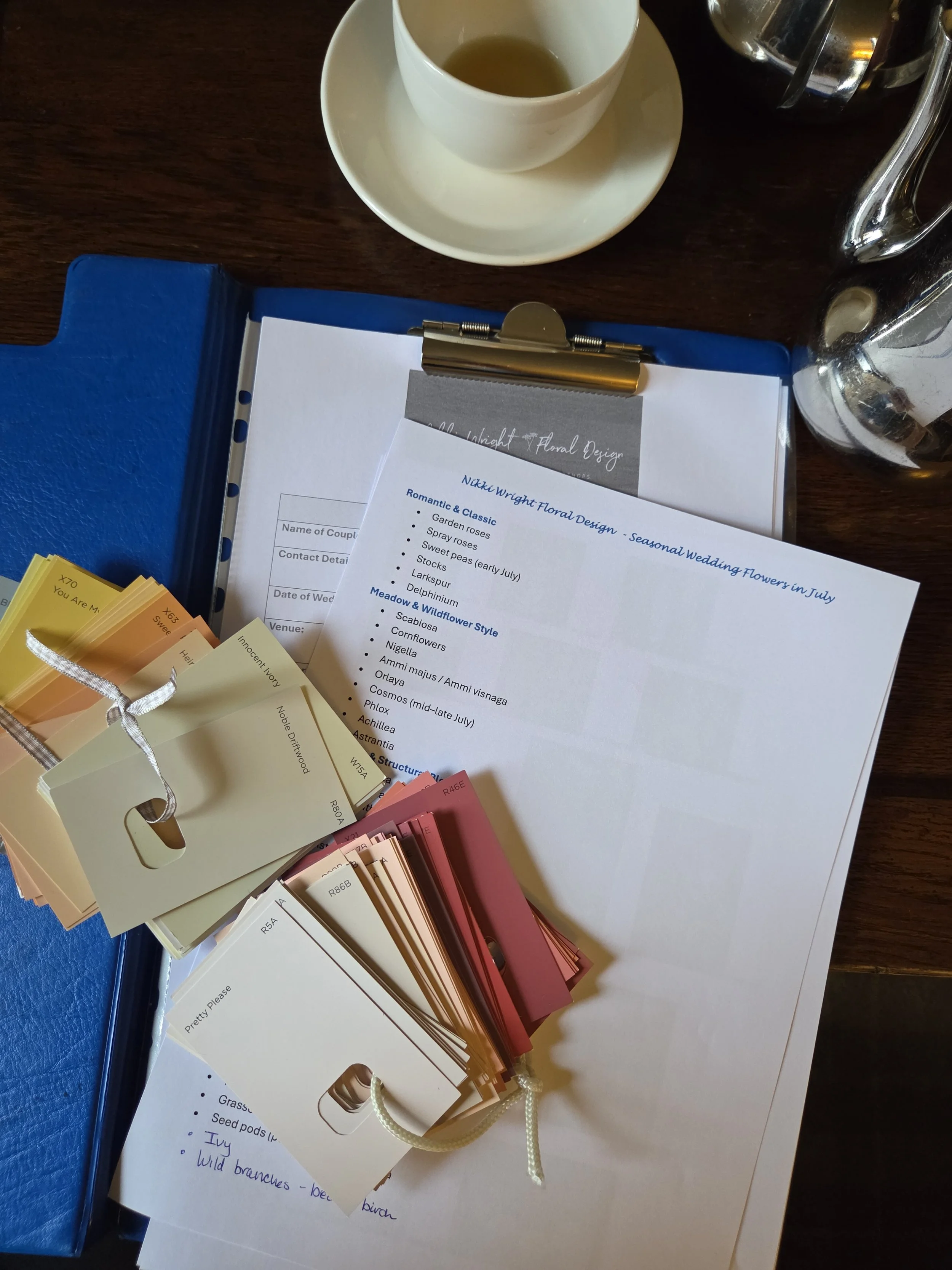 Color swatches and fabric samples for floral and garden design display on a table, alongside a white sheet of paper with handwritten notes and a tea cup nearby.