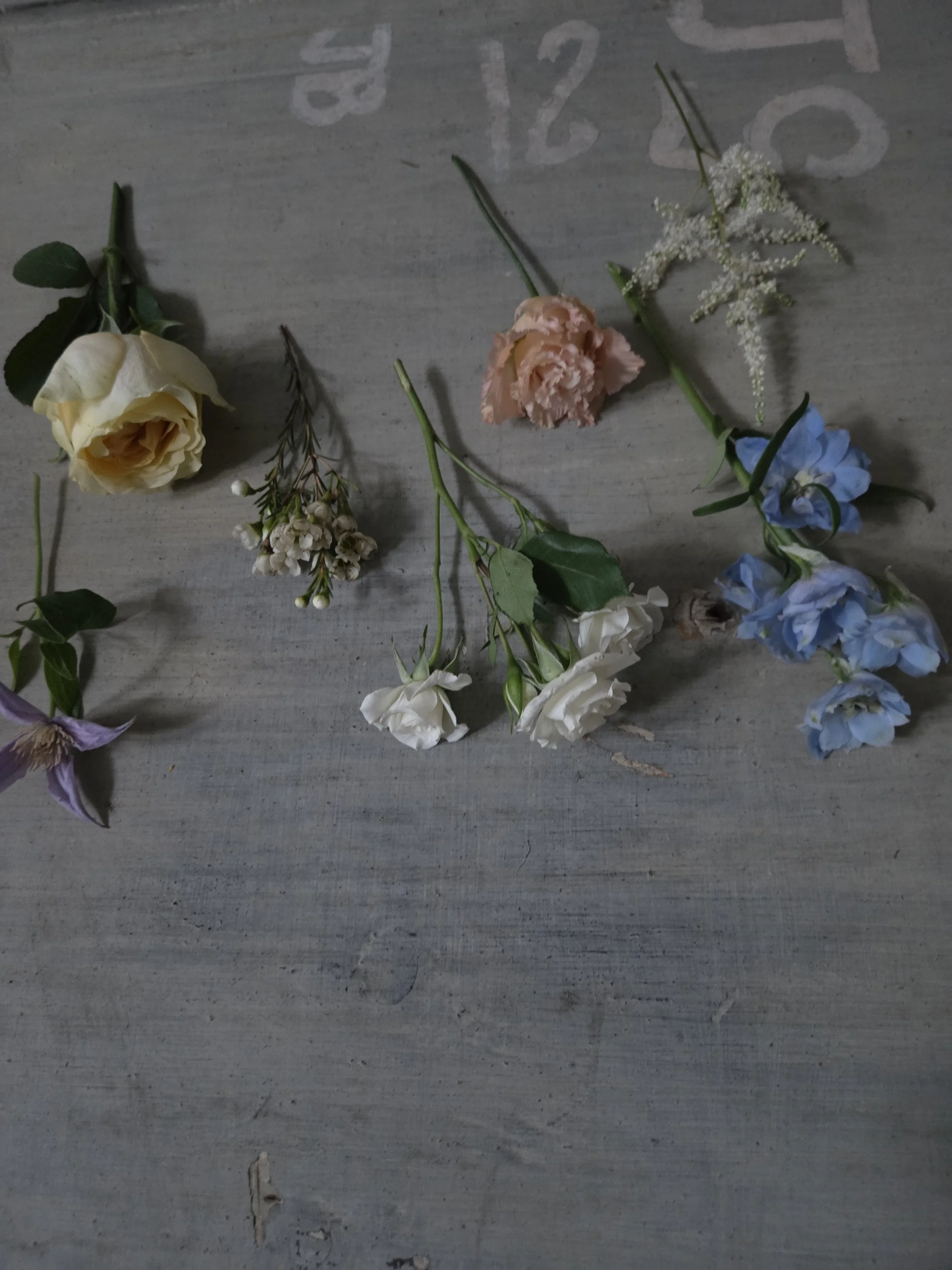 Various small flowers and sprigs of greenery spread on a wooden surface, including a yellow rose, white flowers, pink, blue, white, and lavender blooms.