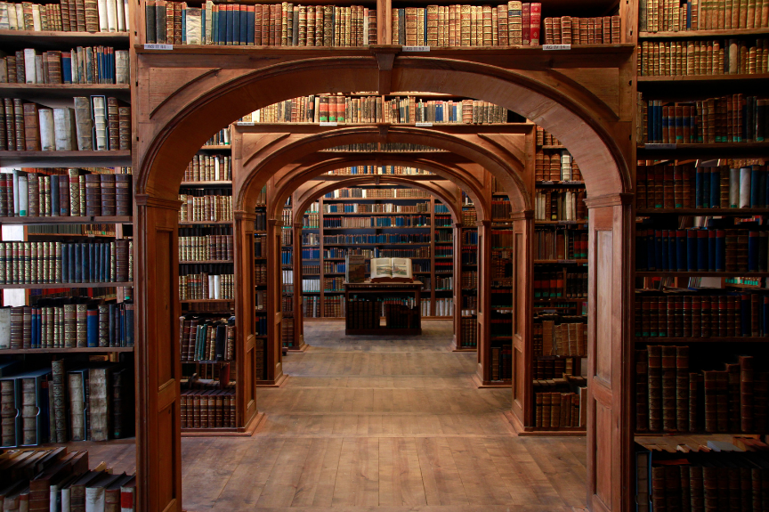 Une bibliothèque ancienne avec des étagères en bois remplies de livres, des arches en bois sculpté formant un couloir, et un livre ouvert sur un pupitre au centre.