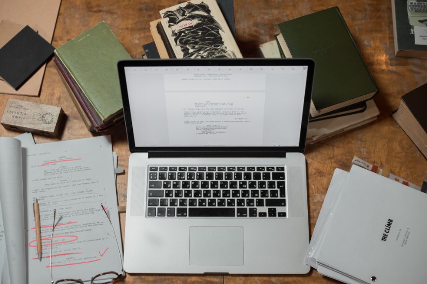 Un ordinateur portable ouvert entouré de livres, papiers, crayons et lunettes sur une table en bois.