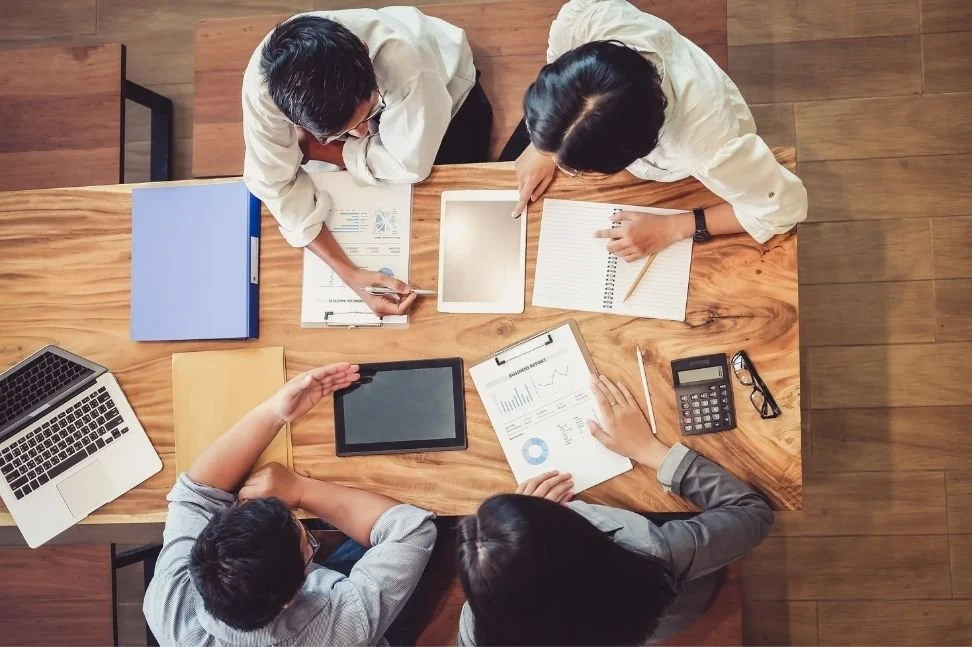 quatre personnes dans une réunion d'affaires autour d'une table en bois, avec des ordinateurs, des tablettes, des documents, une calculatrice et des stylos.