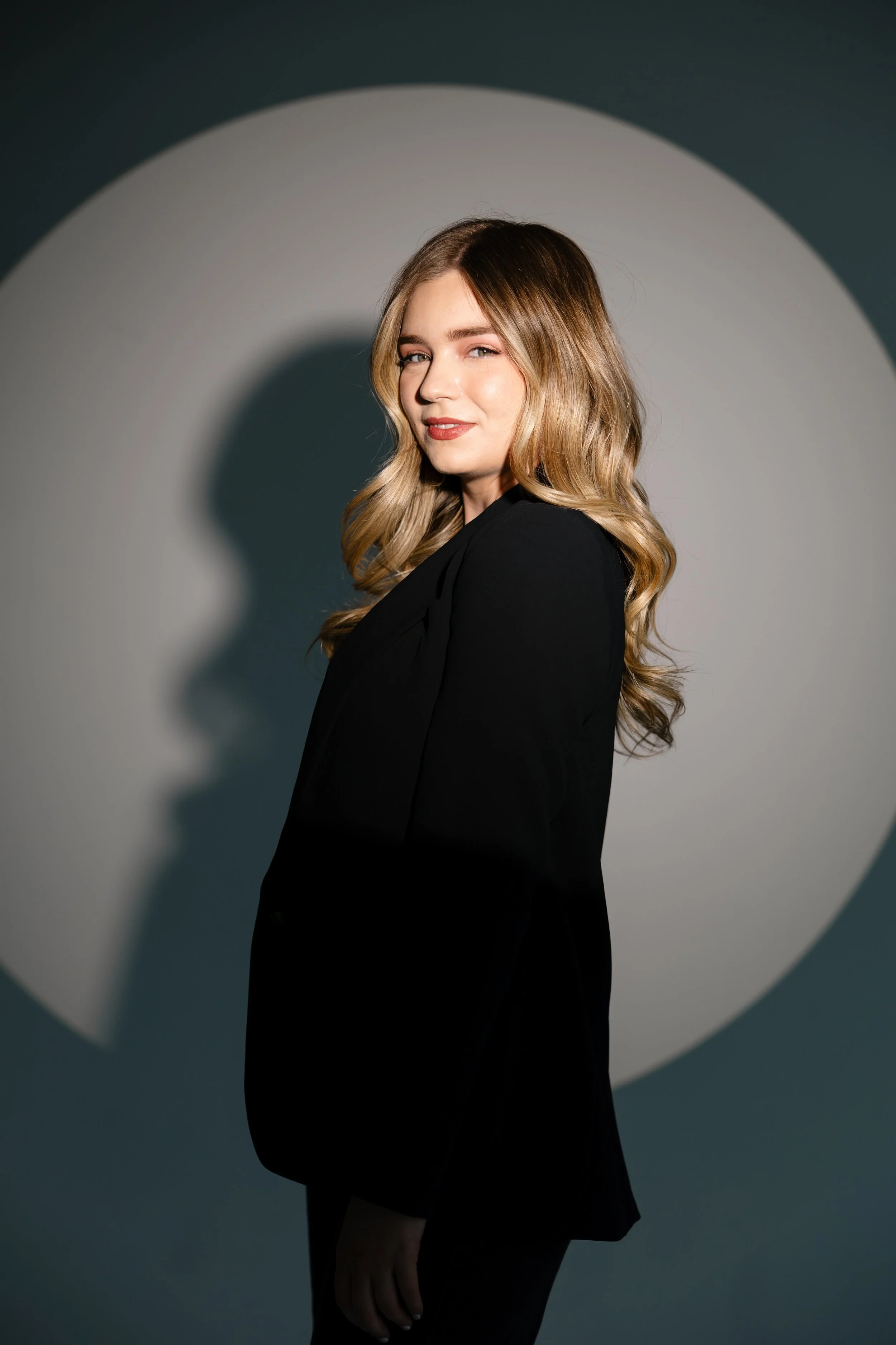 A woman with wavy blonde hair wearing a black blazer, posing against a backdrop with a circular spotlight effect.