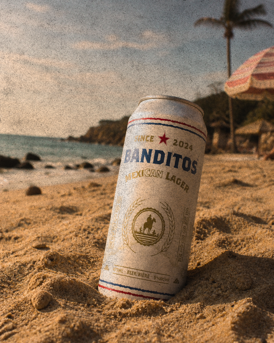 A can of Banditos Mexican lager beer partially buried in sandy beach with a beach umbrella, palm tree, and ocean in the background during sunset.