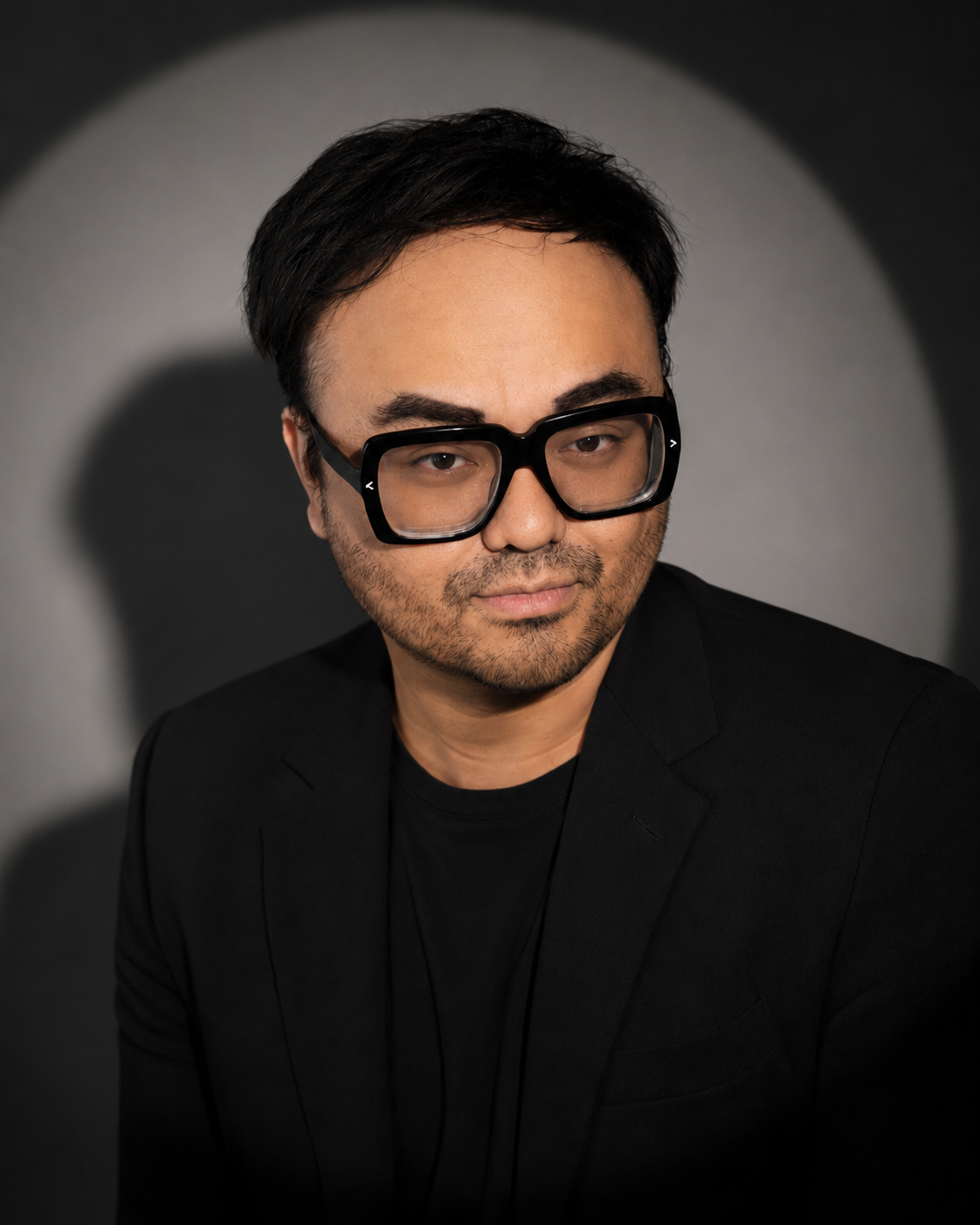 A man with dark hair, wearing oversized black glasses and a black blazer, posed against a dark background with a circular spotlight.