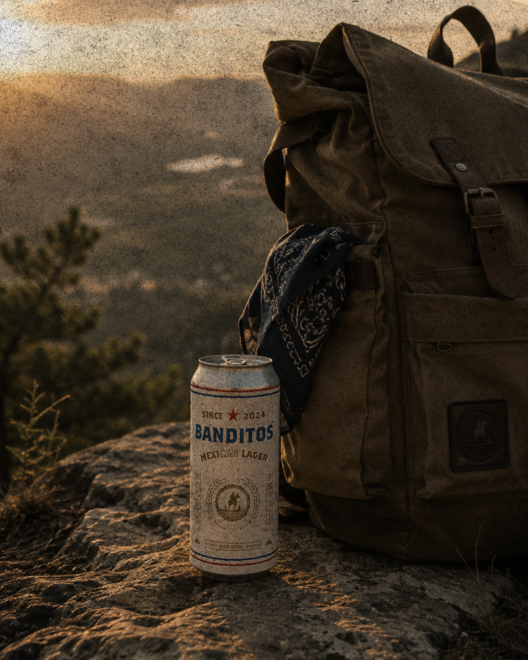 A can of Banditos Mexican Lager next to a brown backpack with a bandana attached, on a rocky surface during sunset with mountains and a tree in the background.