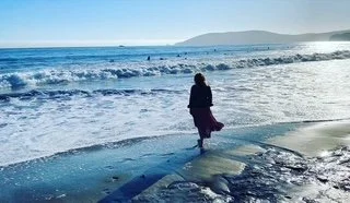 A person standing on the beach facing the ocean with waves and a distant landmass in the background.