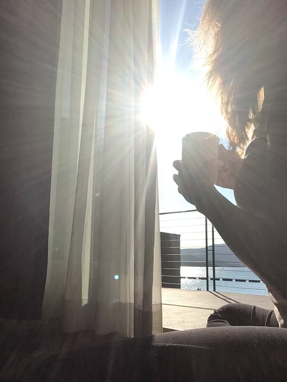 Person sitting near a window, drinking coffee, with sunlight shining brightly through the window, casting rays and illuminating sheer curtains, with a view of water and mountains outside.