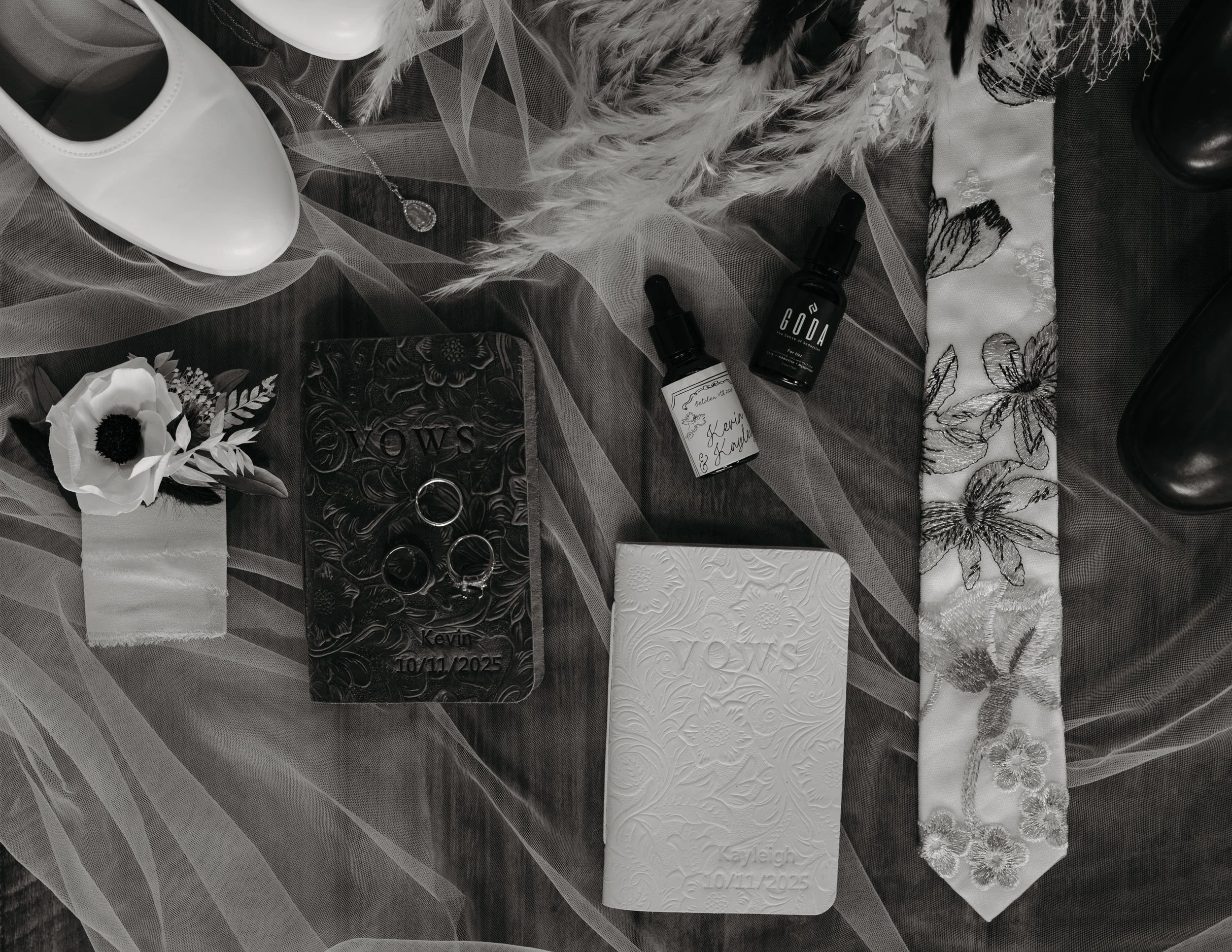 A wedding flat lay with a white shoe, white flower, jewelry, essential oils, black boots, a decorative blanket, and engraved white cards on sheer fabric.