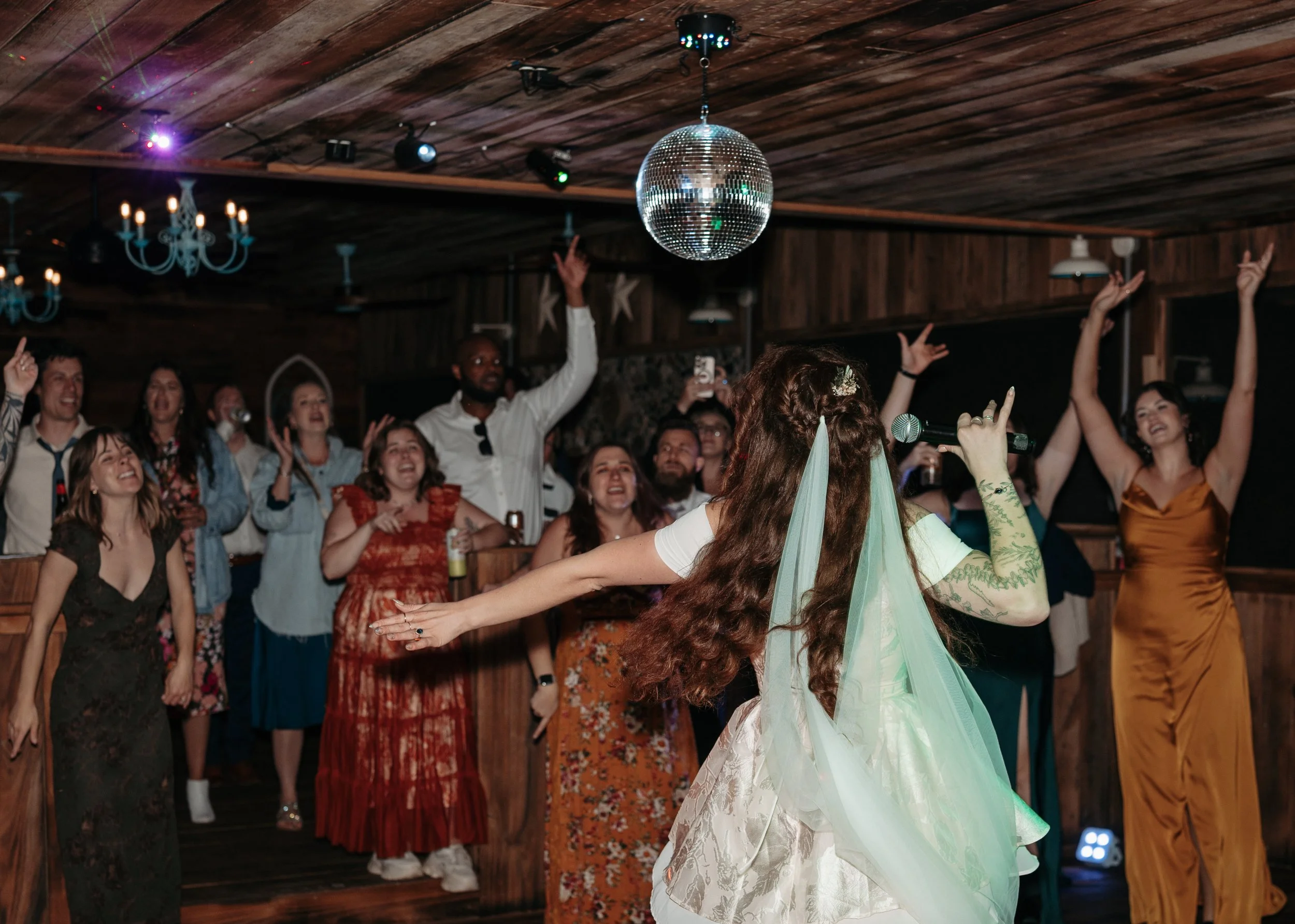 A lively party scene with a group of people dancing and celebrating indoors. A woman with long hair and a light-colored dress is seen from behind, holding a microphone. The room has wooden walls and ceiling, and a large disco ball hangs from the ceiling, reflecting colorful lights. People are smiling, raising their hands, and enjoying the moment.