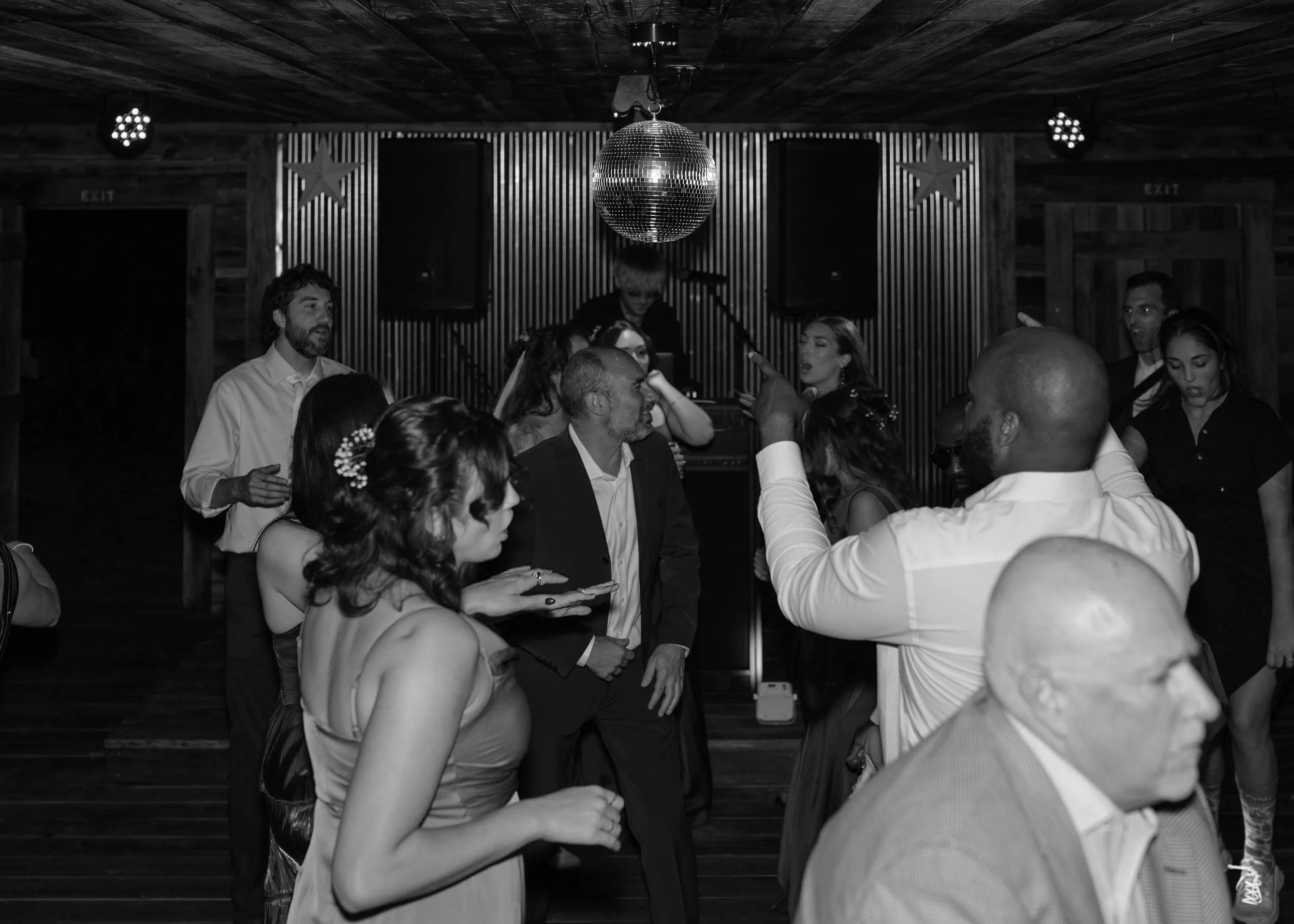 People dancing and socializing at a party with a disco ball hanging from the ceiling, wooden walls, and star decorations.