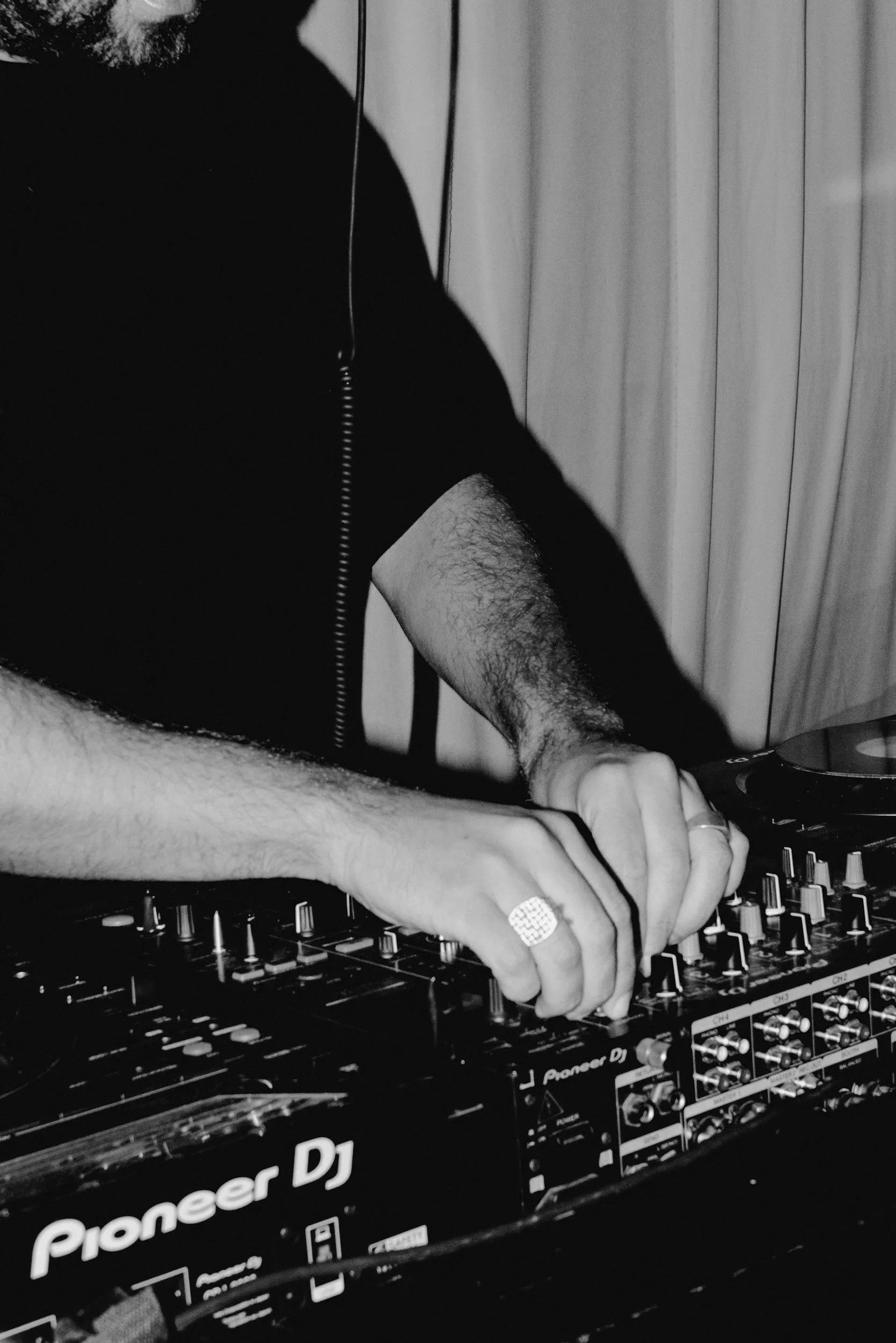A person with a beard wearing a black shirt, with a ring on their finger, is operating a Pioneer DJ mixer or controller.