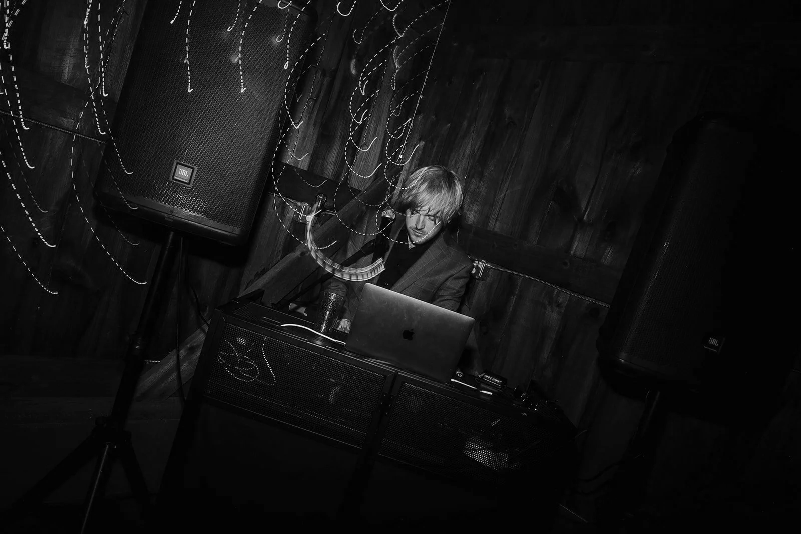 A man DJing in a dimly lit room with wooden walls, using a laptop, with speakers and light trails around him.