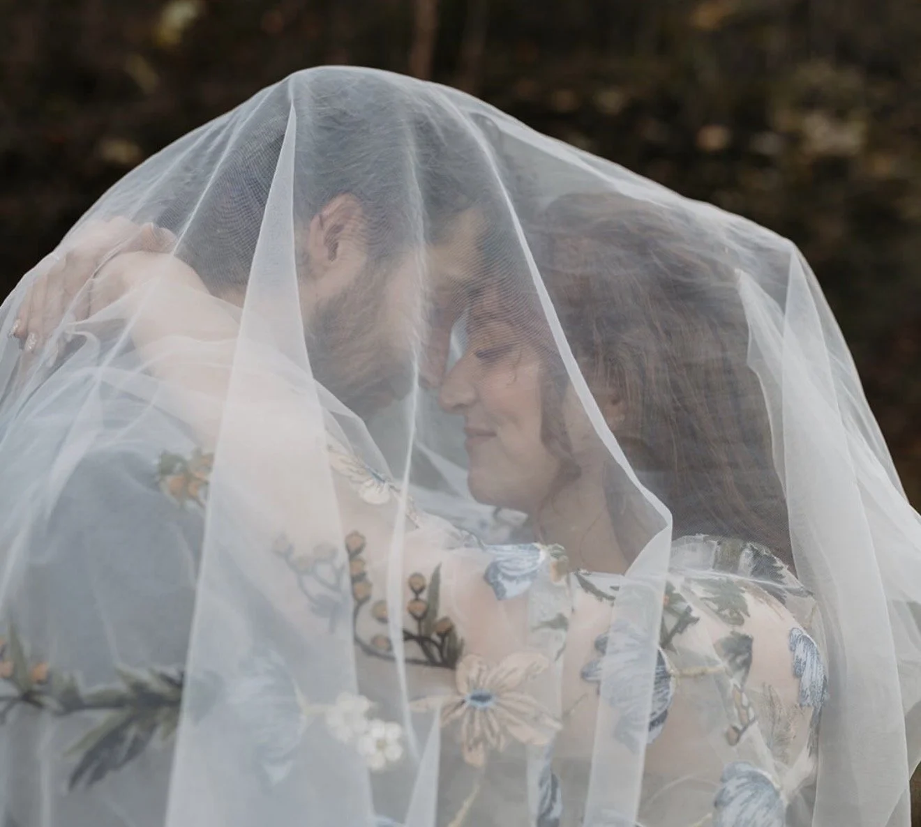 A couple under a wedding veil, touching foreheads and smiling softly.
