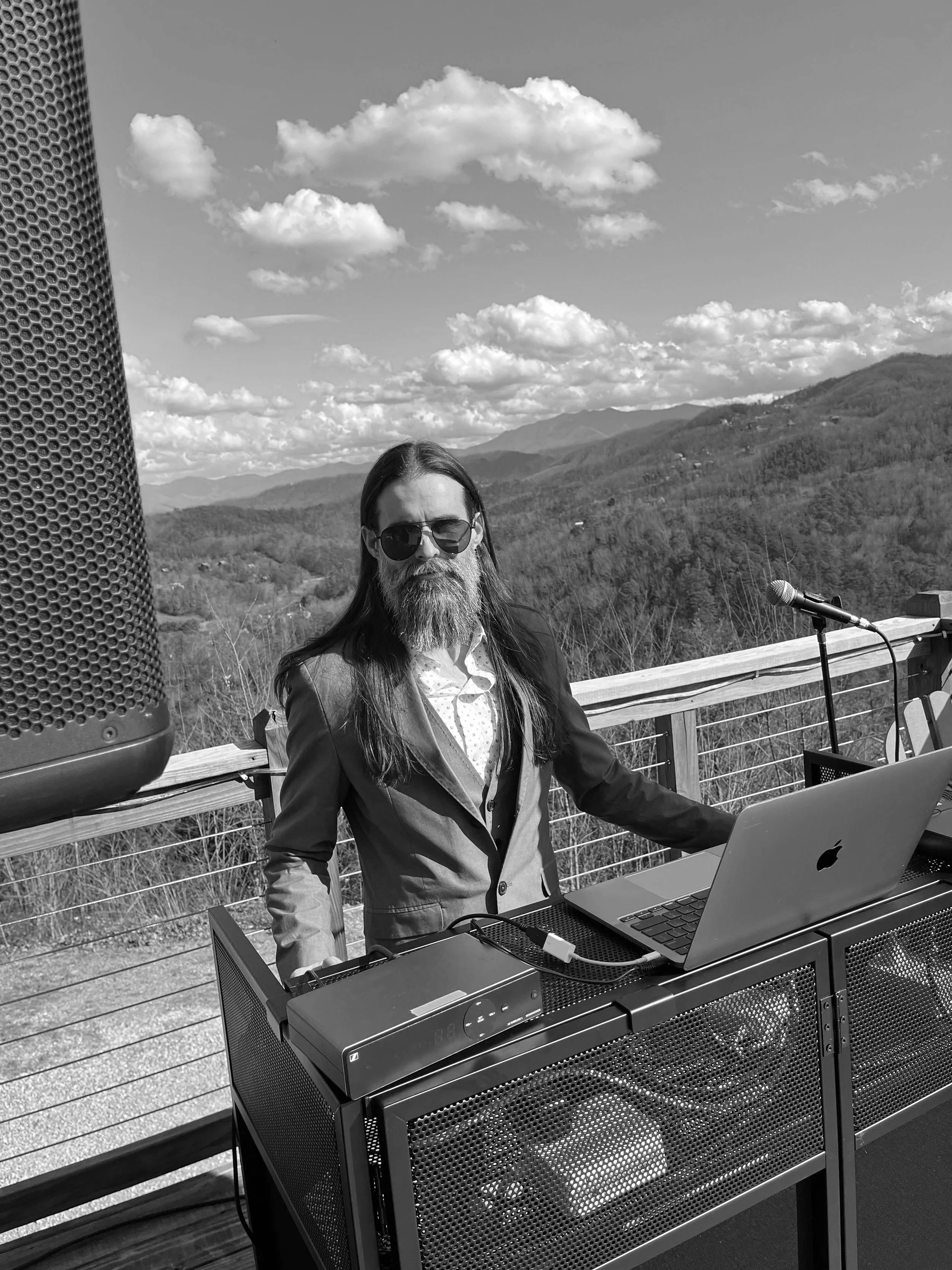 A man with long hair and a beard wearing sunglasses and a blazer, standing at an outdoor table with a laptop, mountain landscape in the background, microphone on a stand in front of him.