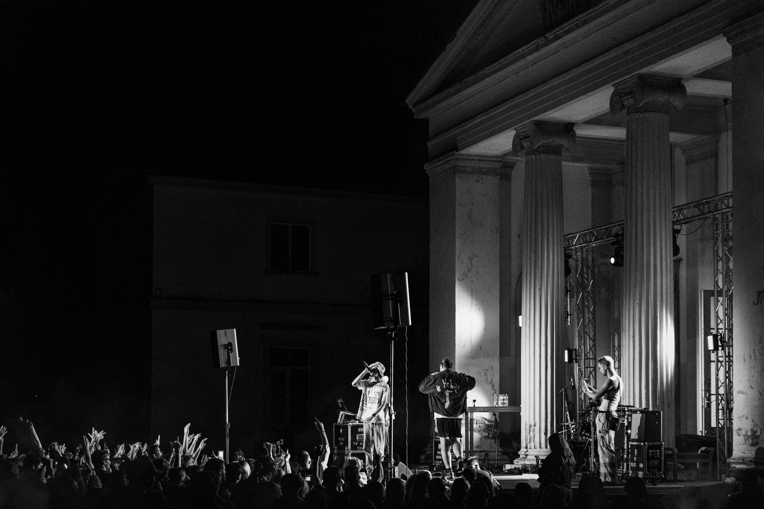 A black and white photo of a live musical performance on a stage with an audience watching. The stage has large classical columns and a high ceiling. Several performers are visible, standing and playing instruments or microphones, with band equipment around them. Audience members are reaching towards the stage with raised hands.