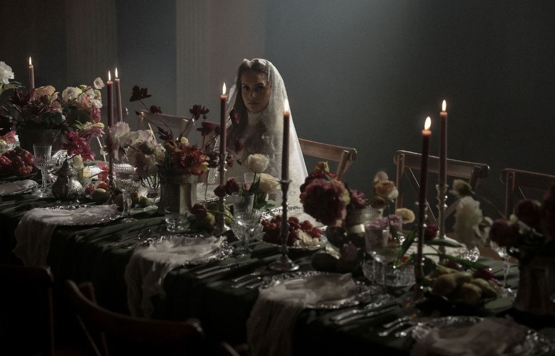 A woman wearing a white bridal veil sits at a decorated dinner table with floral arrangements, candles, and glassware in a dimly lit room.