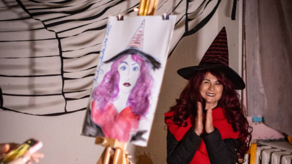 A woman dressed as a witch with curly red hair, wearing a black and red costume and a pointed witch hat, smiling with her hands pressed together. An easel with a painting of a woman with pink hair and a witch hat is in the foreground, and a decorative wall with black abstract lines is in the background.