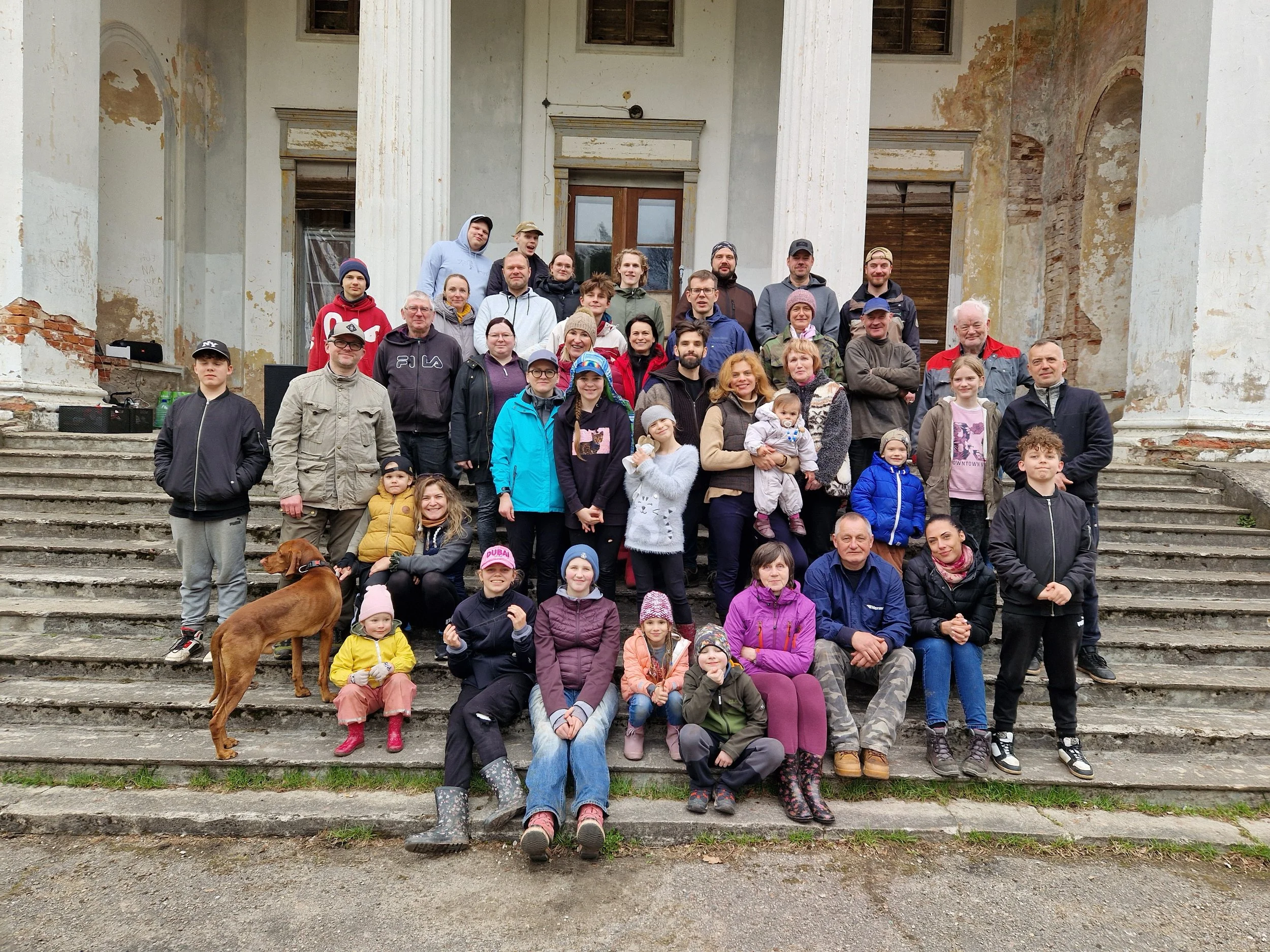 A large group of people, including adults and children, posing on the steps of a weathered, old building with large columns. The building appears to be in a state of disrepair with peeling paint and exposed bricks. The group is smiling and appears happy, with some children sitting on the stairs and adults standing behind them.