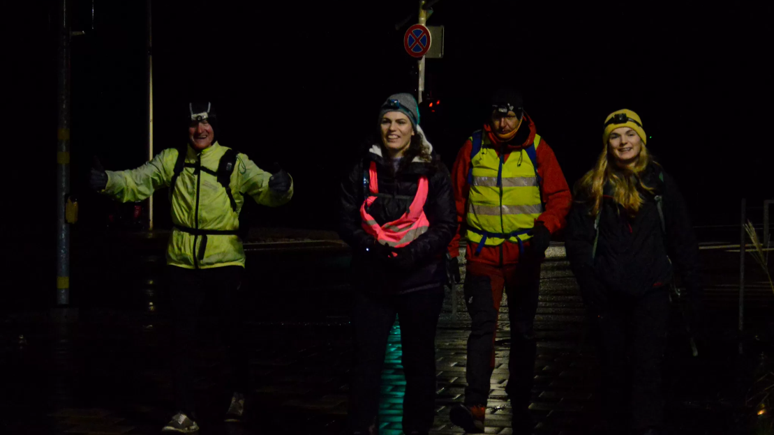 Four people in outdoor gear walking at night, smiling, on wet pavement, with street signs behind them.