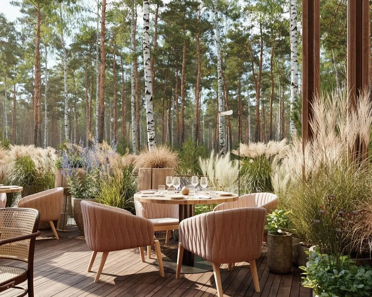 Restaurant patio with pink upholstered chairs, set with wine glasses and a centerpiece, overlooking a forest with birch and pine trees, surrounded by tall grasses and potted plants.