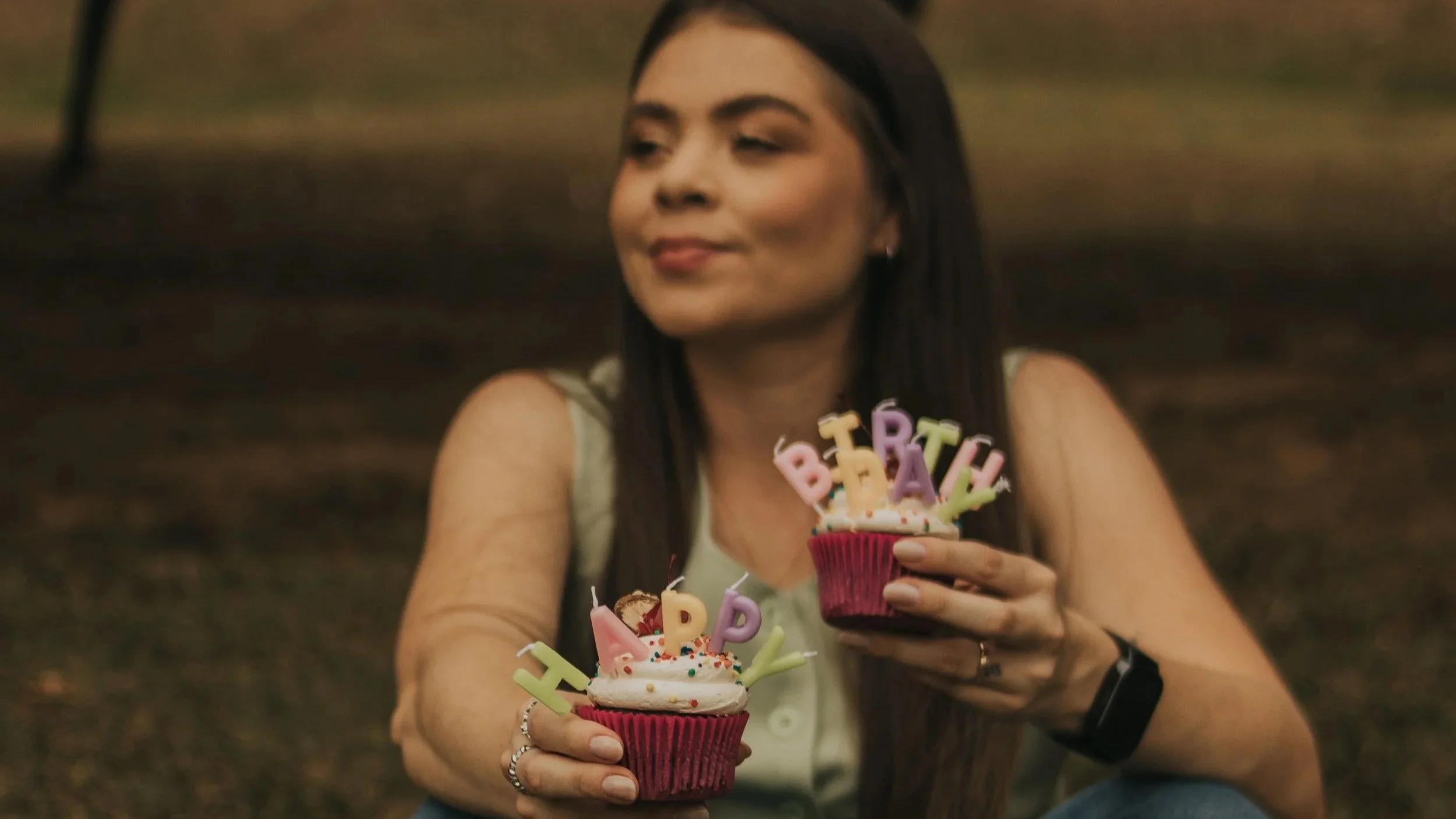 A woman with long dark hair holding birthday cupcakes with colorful "HAPPY BIRTHDAY" candles, smiling softly in an outdoor setting.